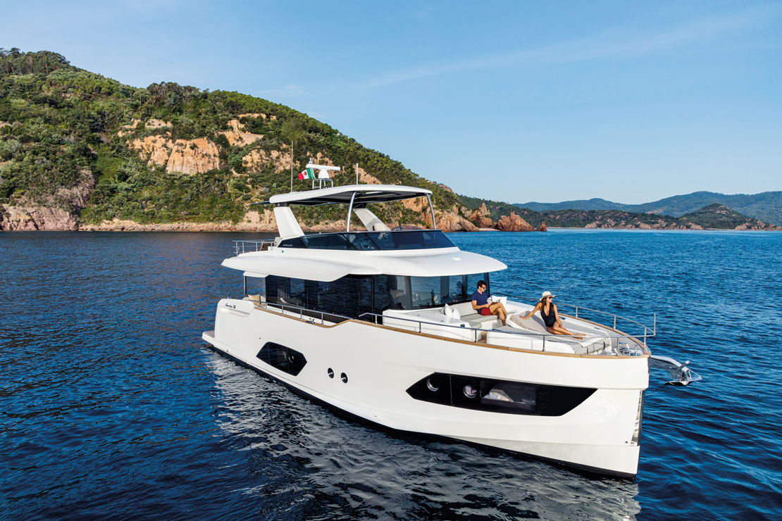The Navetta 58 at rest close inshore, guests on the foredeck and a wooded headland behind.