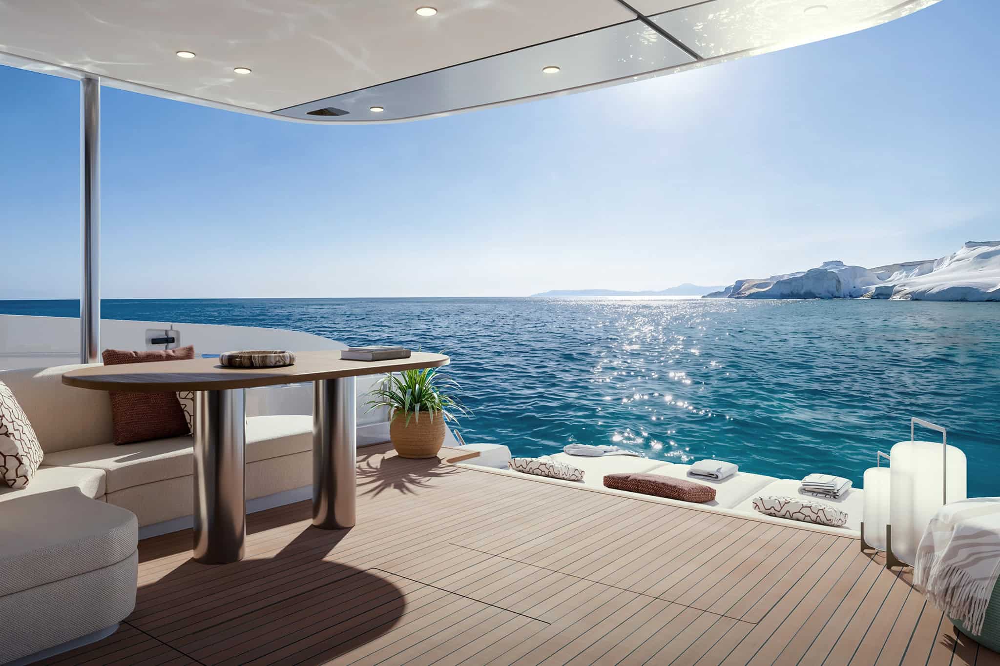 The Terrace Cockpit looking aft over the sea, an oval dining table and bench seating on teak under the deck above, with sunpads at the open stern and a cliff coastline in the distance
