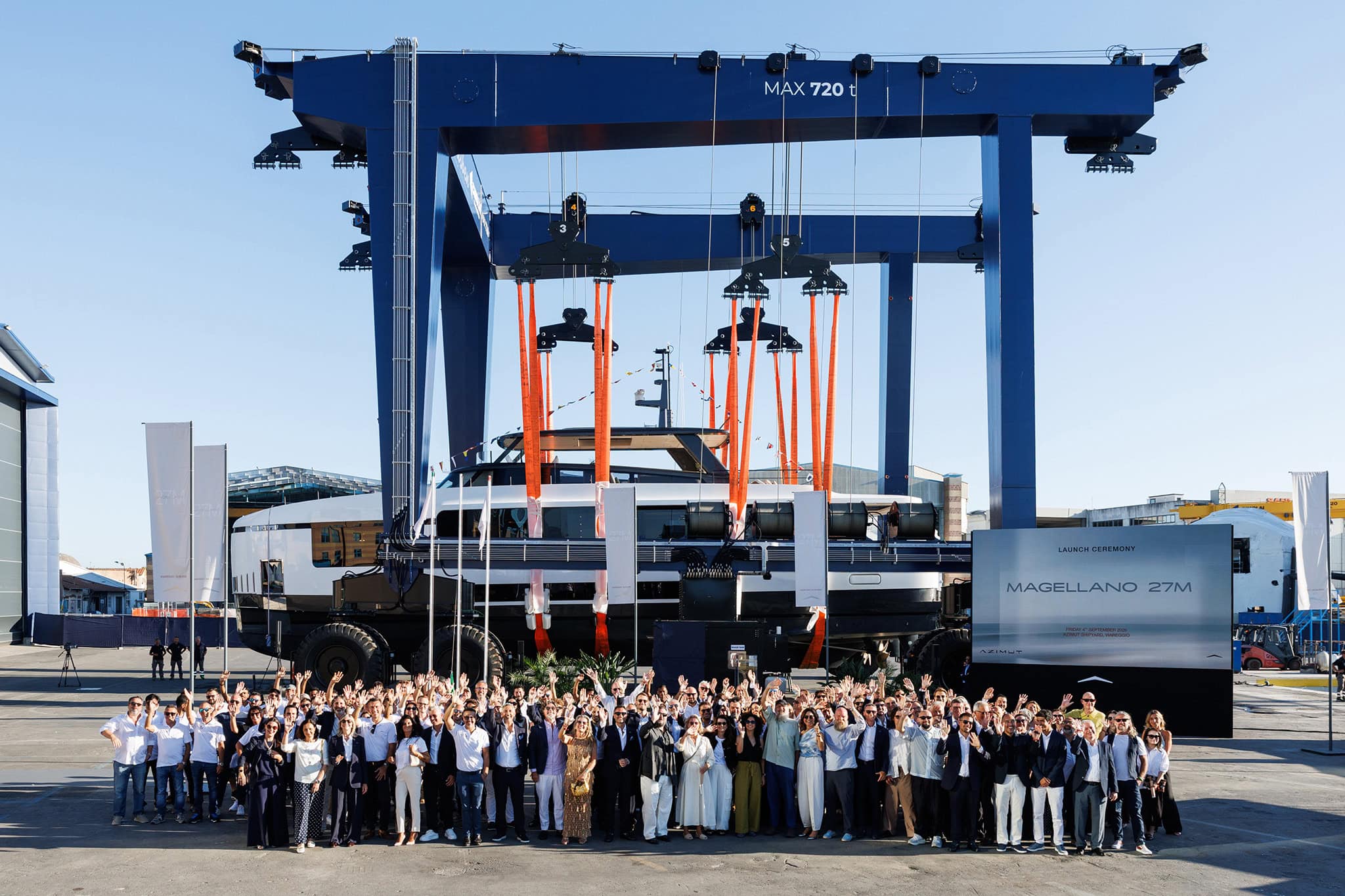 The Magellano 27M held in a blue travel lift at the Azimut yard in Viareggio with a large crowd of staff and guests waving in front of her, beside a board reading Launch Ceremony Magellano 27M