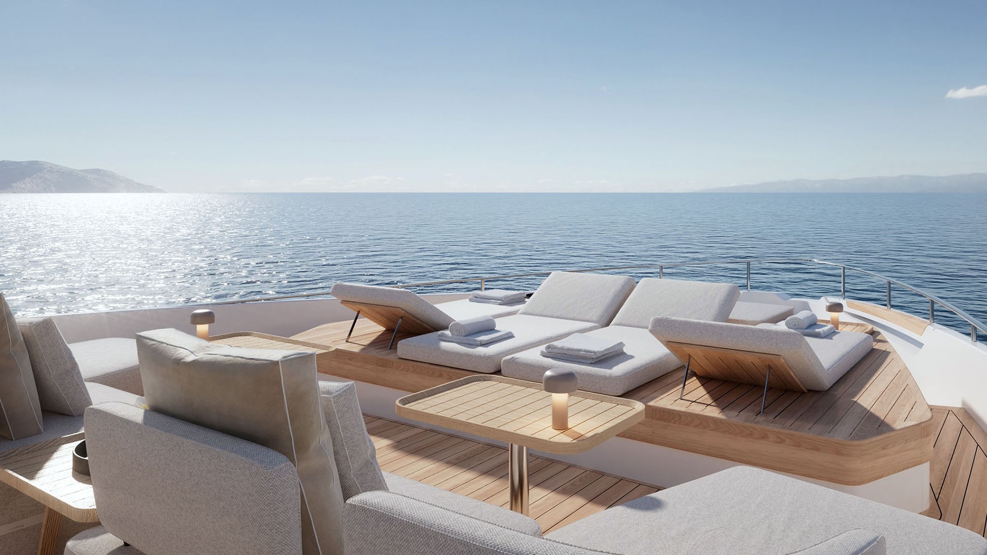 The foredeck seating in bright sun, a pair of double sunbeds with rolled towels beyond a group of armchairs and low tables, the sea flat to the horizon