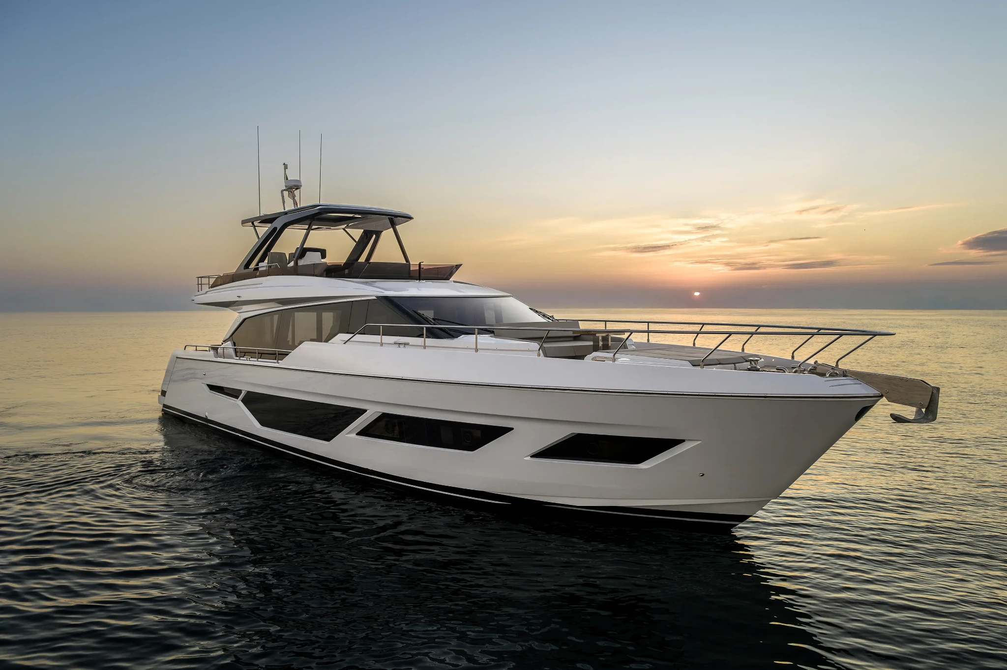 The Ferretti Yachts 720 at anchor in flat calm at sunset, seen from ahead and to one side with the sun setting behind her and the anchor pulpit extended