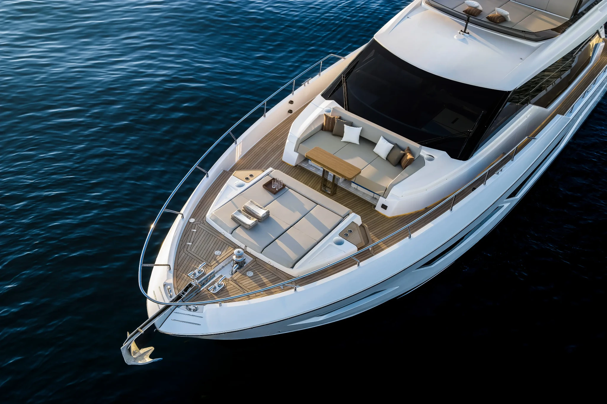 Looking down on the foredeck from above and forward, a large sunpad and a seating group with a folding table set into teak, the anchor and windlass at the stem
