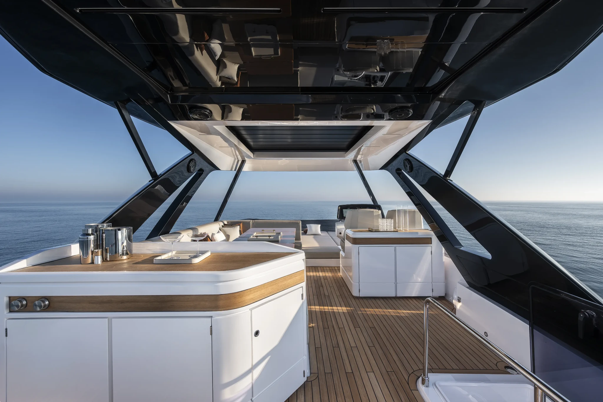 The flybridge looking aft under its dark hardtop, a wet bar with cocktail shakers and glasses in the foreground and a sunpad and seating beyond, open sea on both sides