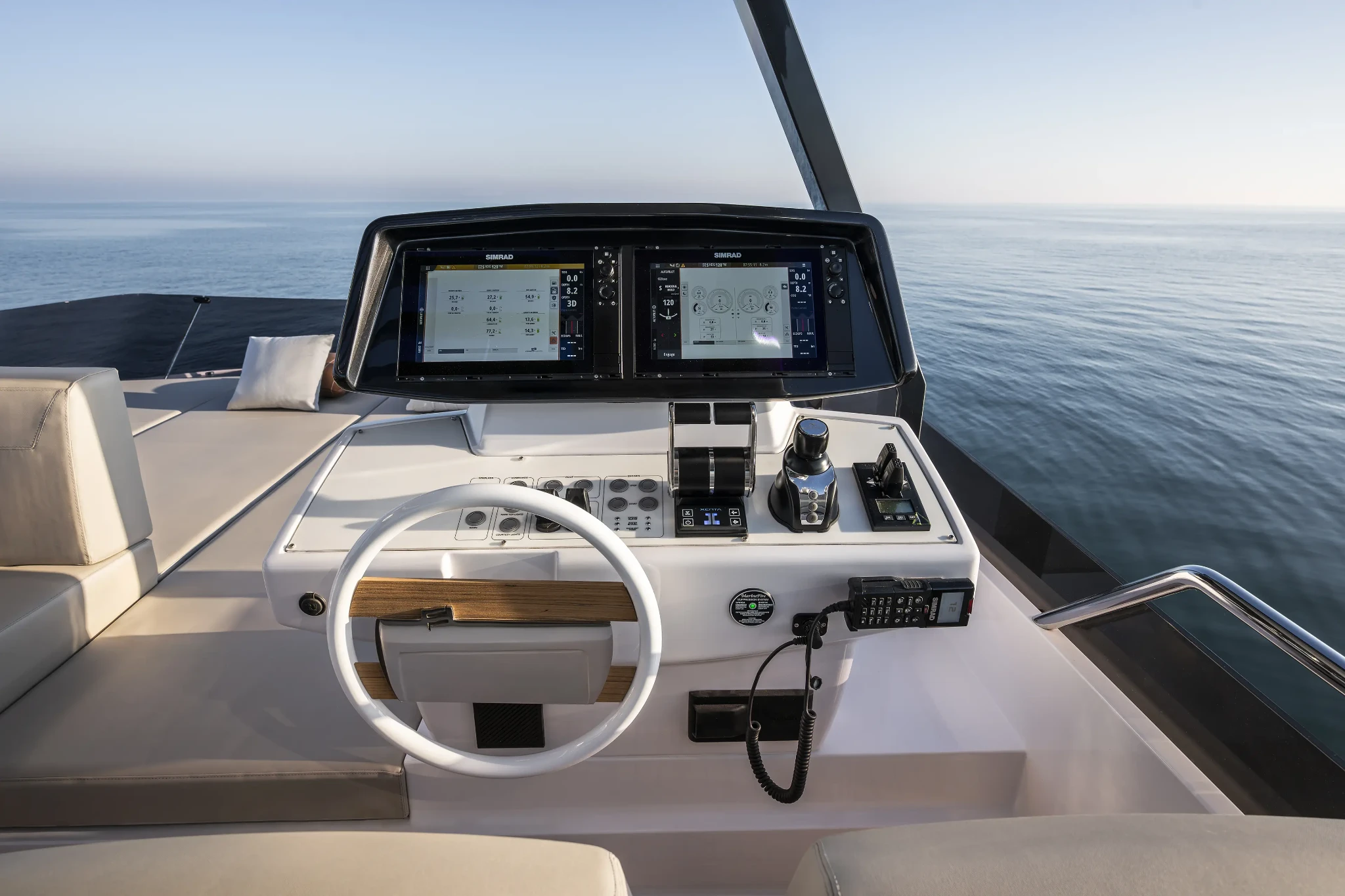 The flybridge helm, a white wheel with a wooden grip below twin Simrad screens, throttles and a joystick on a white console, with the sea beyond