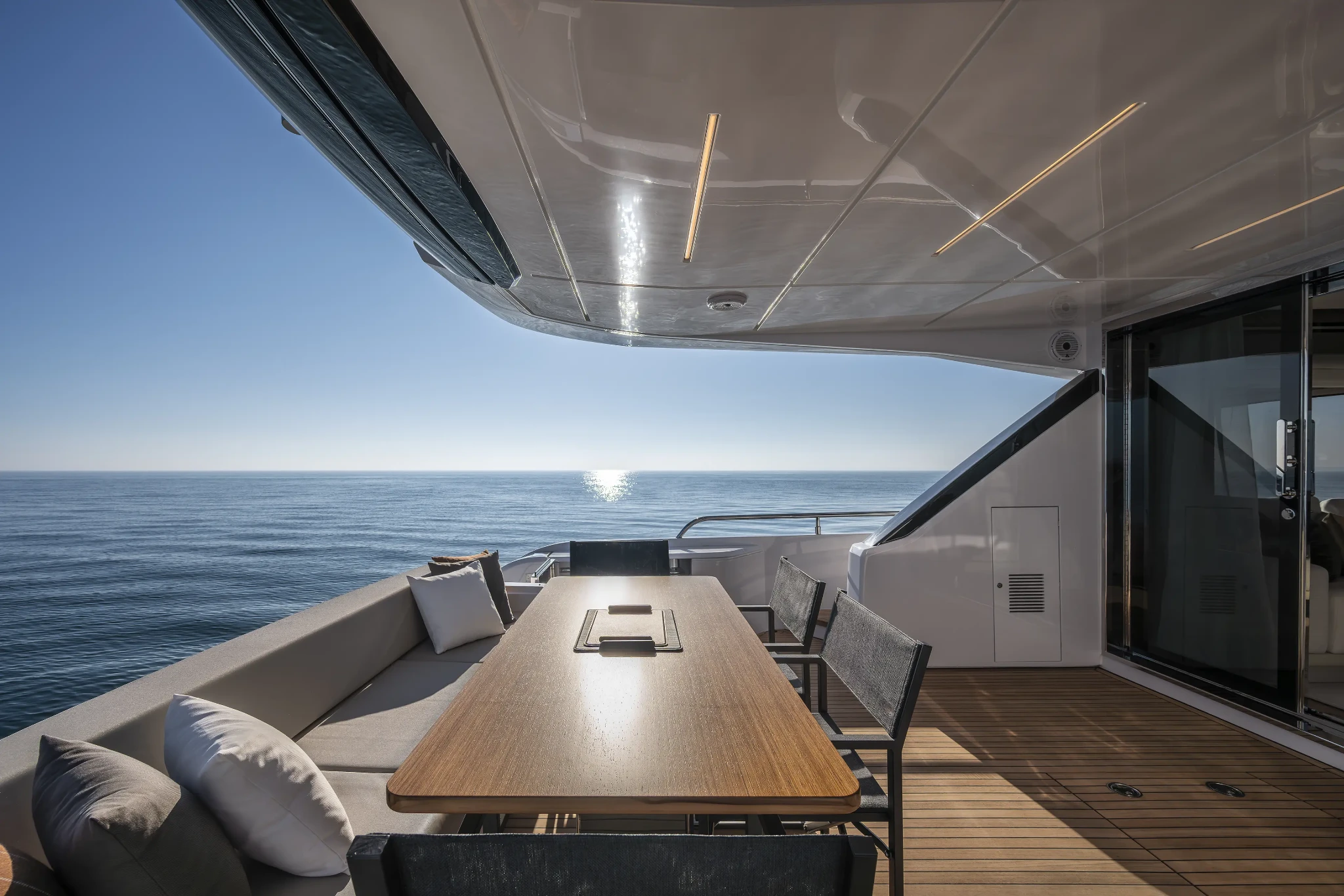 The aft deck dining table in teak with a long bench seat to one side and two chairs opposite, the sea and a low sun filling the open side and glass doors to the saloon at right
