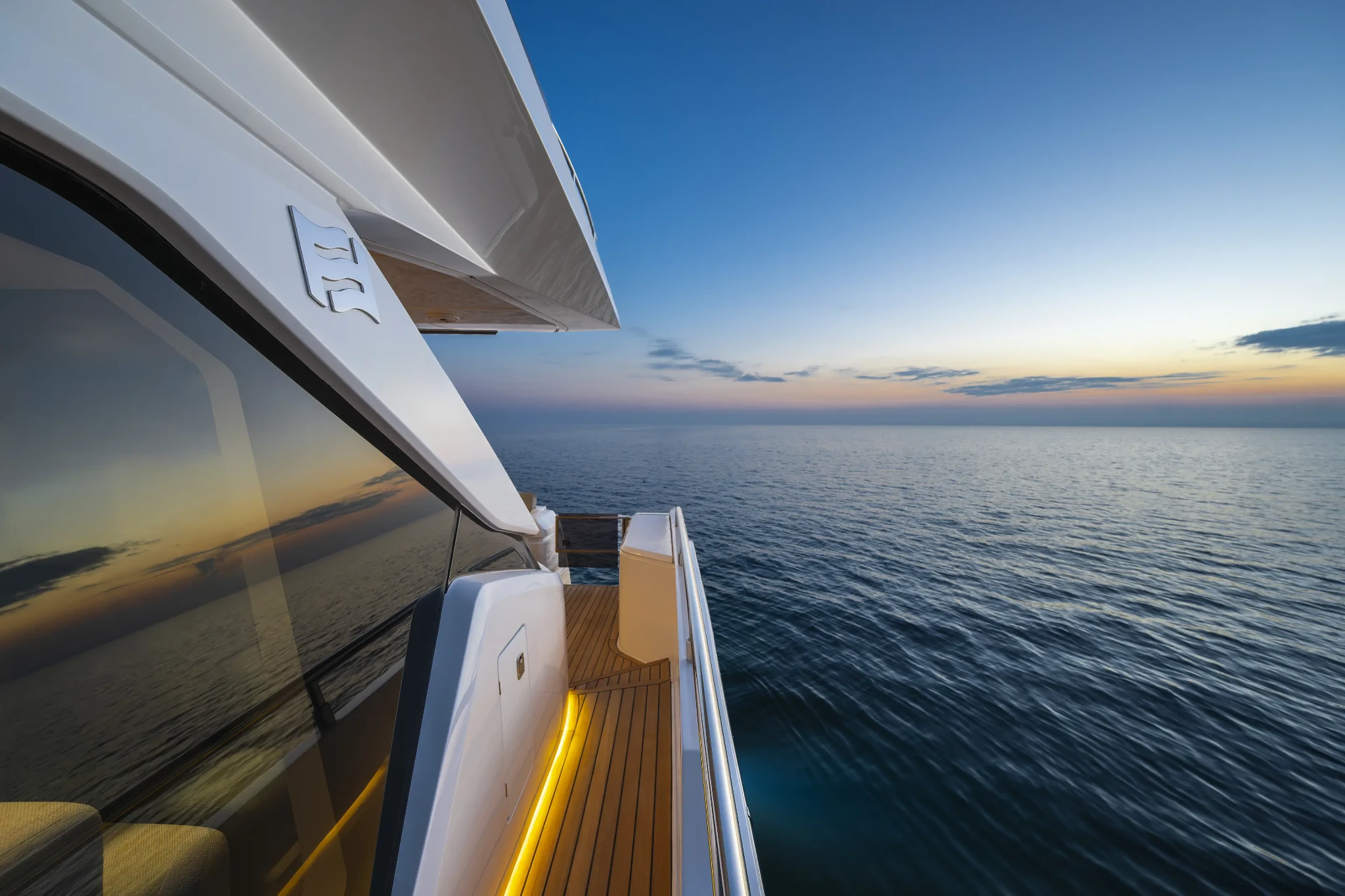 Looking aft along the side deck at dusk, warm strip lighting picking out the teak, with the saloon glazing to the left and the sea to the right