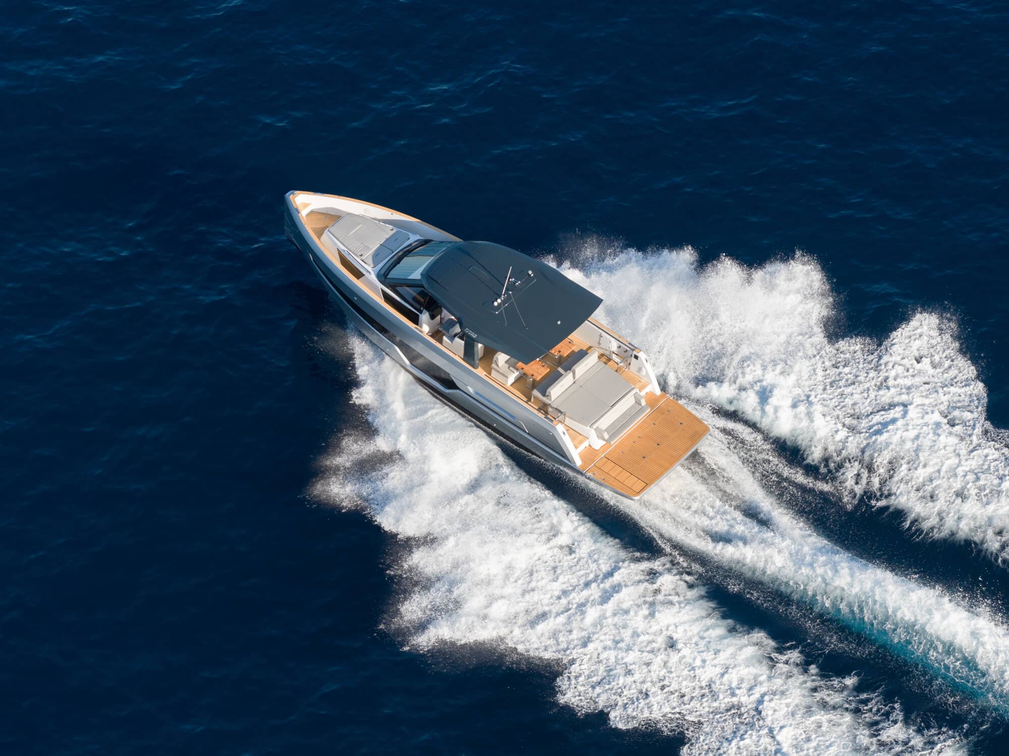 The FJORD 440 photographed from above and astern at speed, twin wakes fanning out white behind her across dark blue water, the cockpit seating and sunpad open to the sky