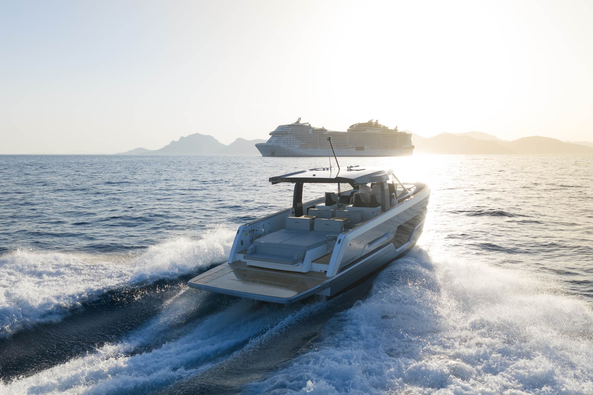 The FJORD 440 seen from astern in evening light with a large cruise ship on the horizon behind her, the aft sunpad, helm seats and hardtop laid out along her length
