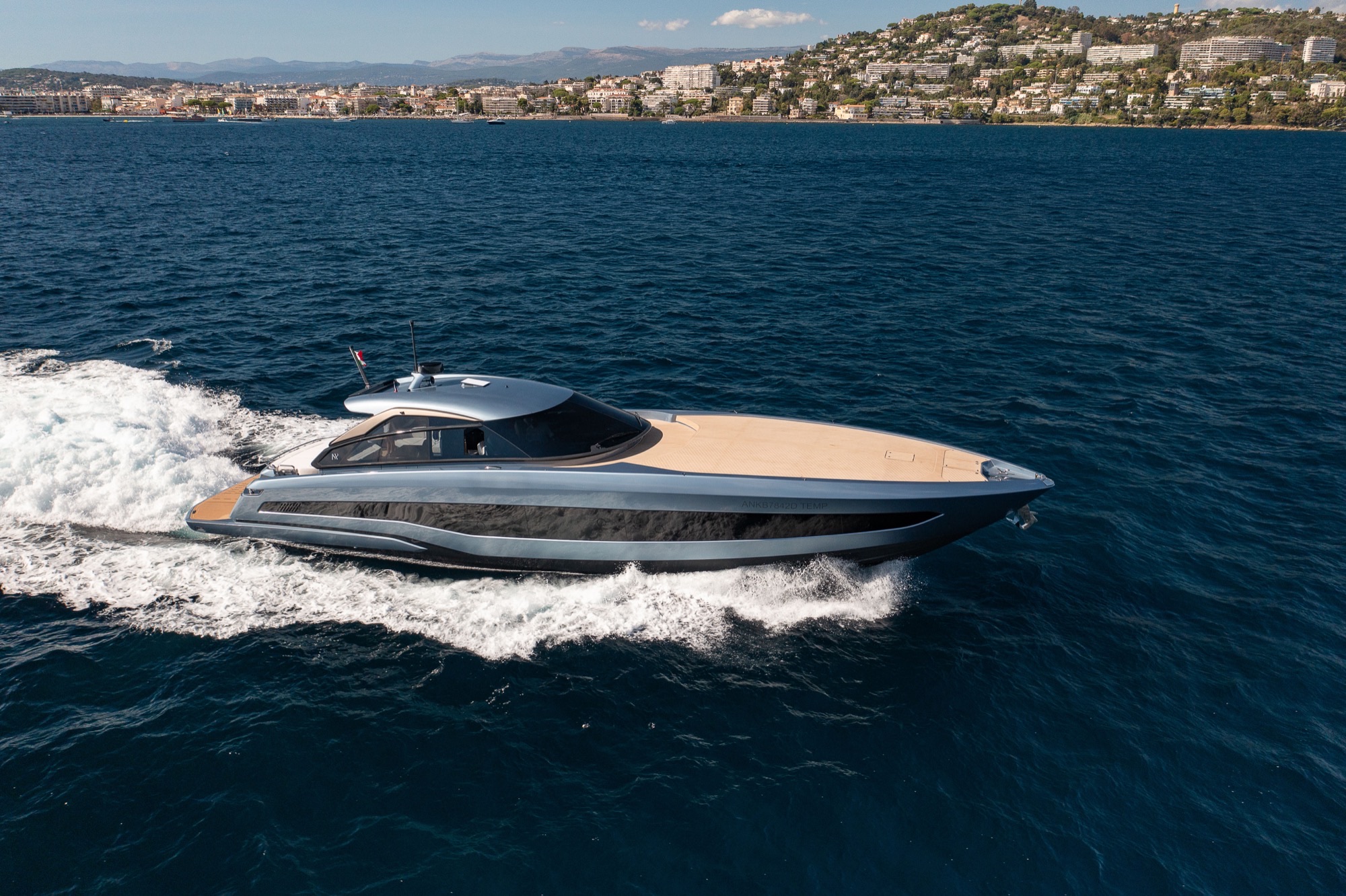 The GTO 70 running past a hillside town, its teak foredeck and silver hull seen from above