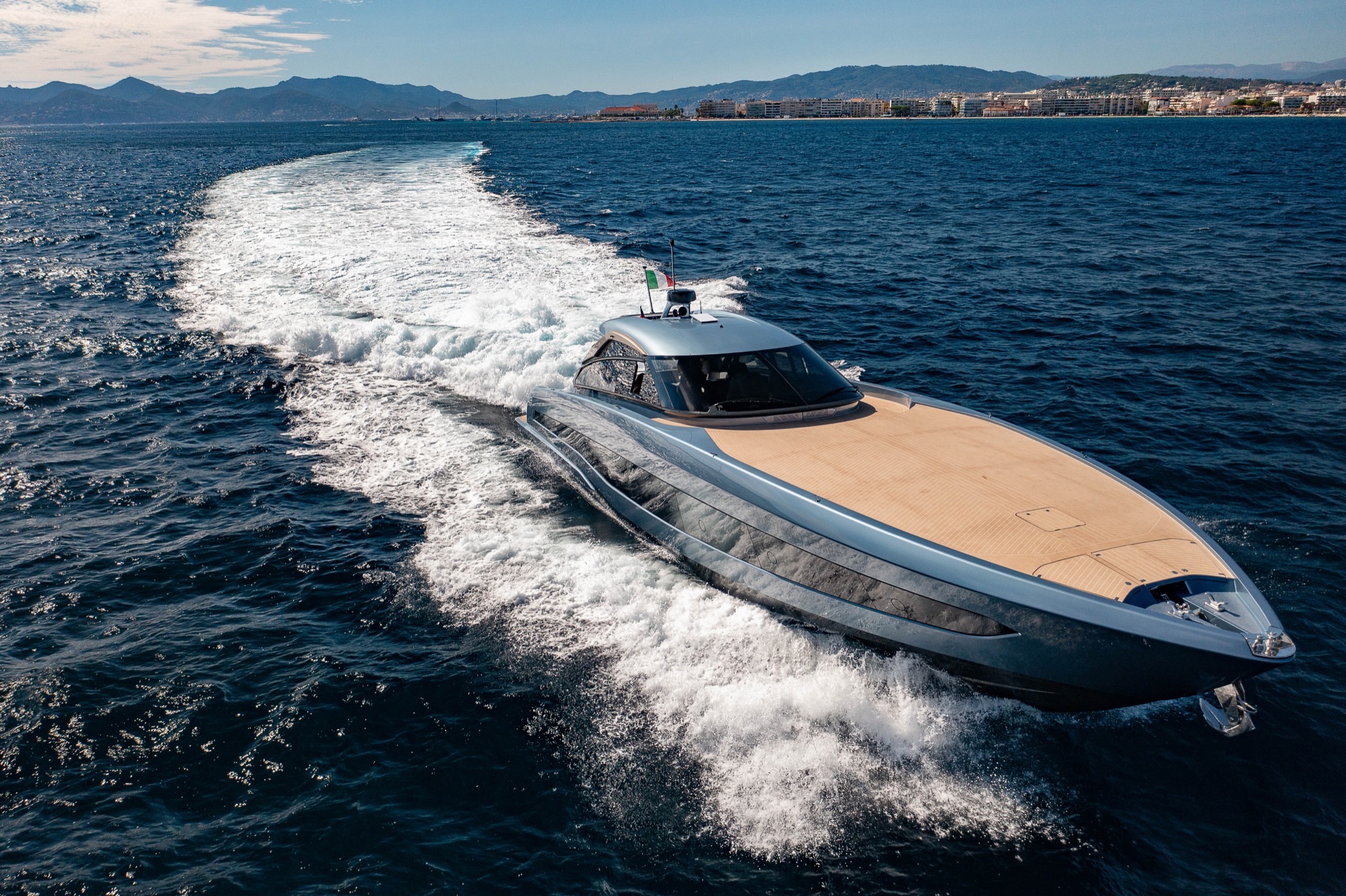 The GTO 70 from the bow quarter, a long wake curving away behind it towards the shore
