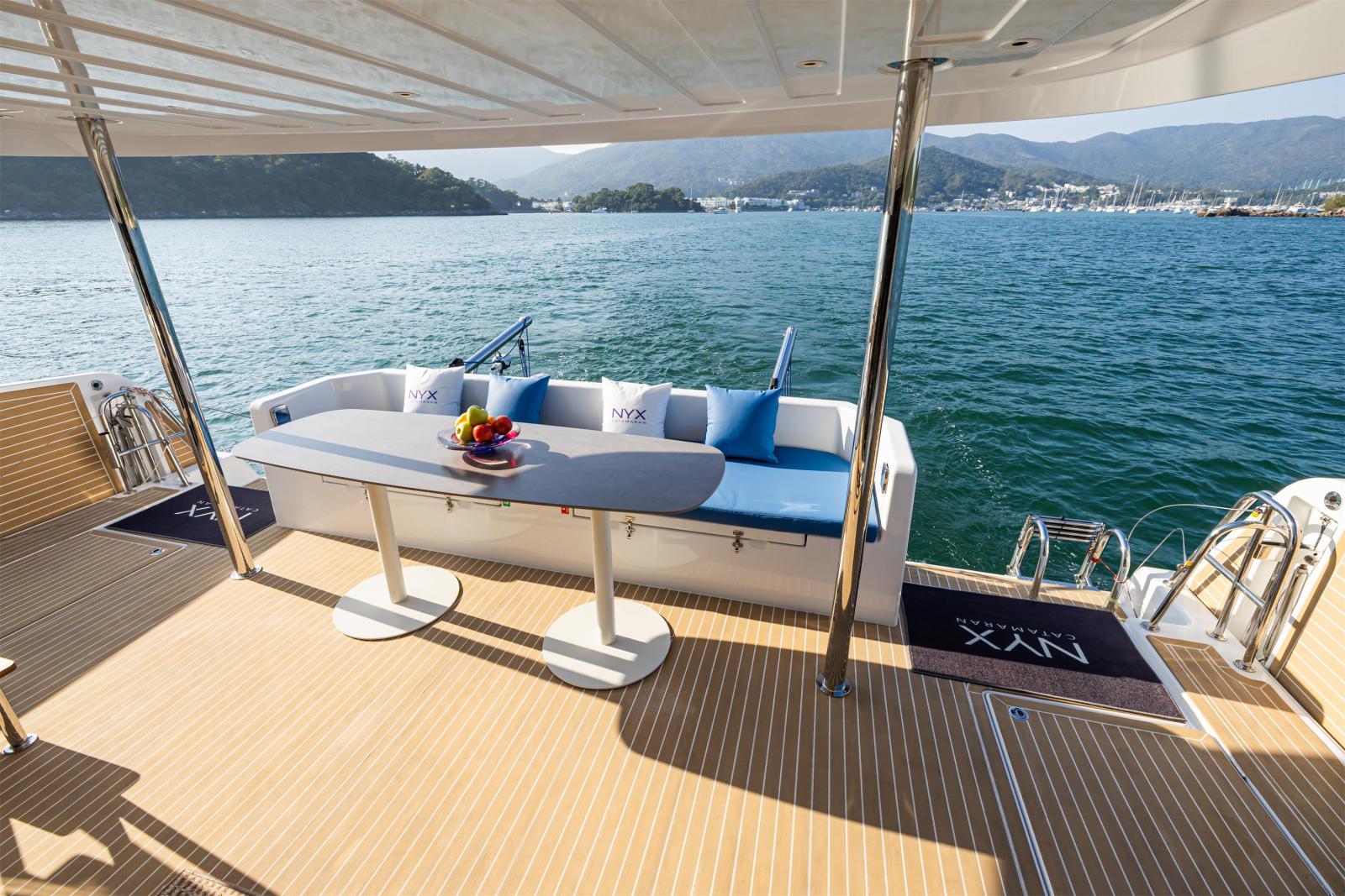 The aft cockpit, with a settee and NYX cushions behind a dining table on teak decking, boarding steps at the transoms and a bay with hills beyond