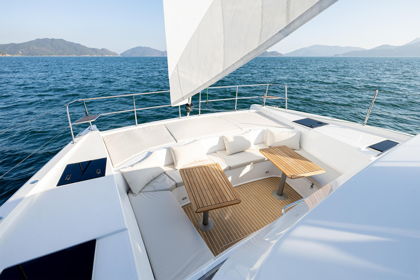 The foredeck lounge between the bows, a cushioned seating area with two teak tables, seen from the coachroof with the jib set above and hills beyond