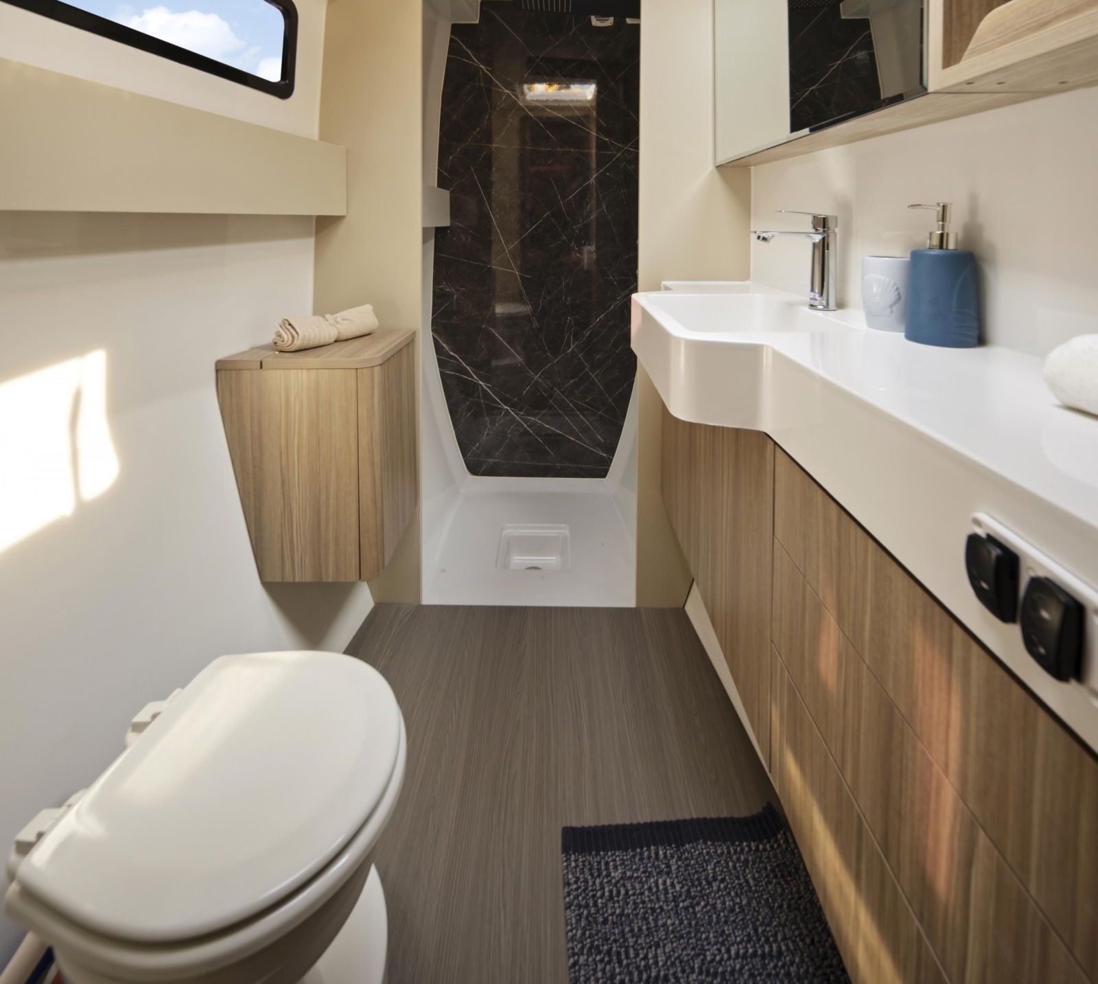 A heads compartment, with a basin on a white worktop, a separate shower stall lined in dark marble-effect panels and a hull window above