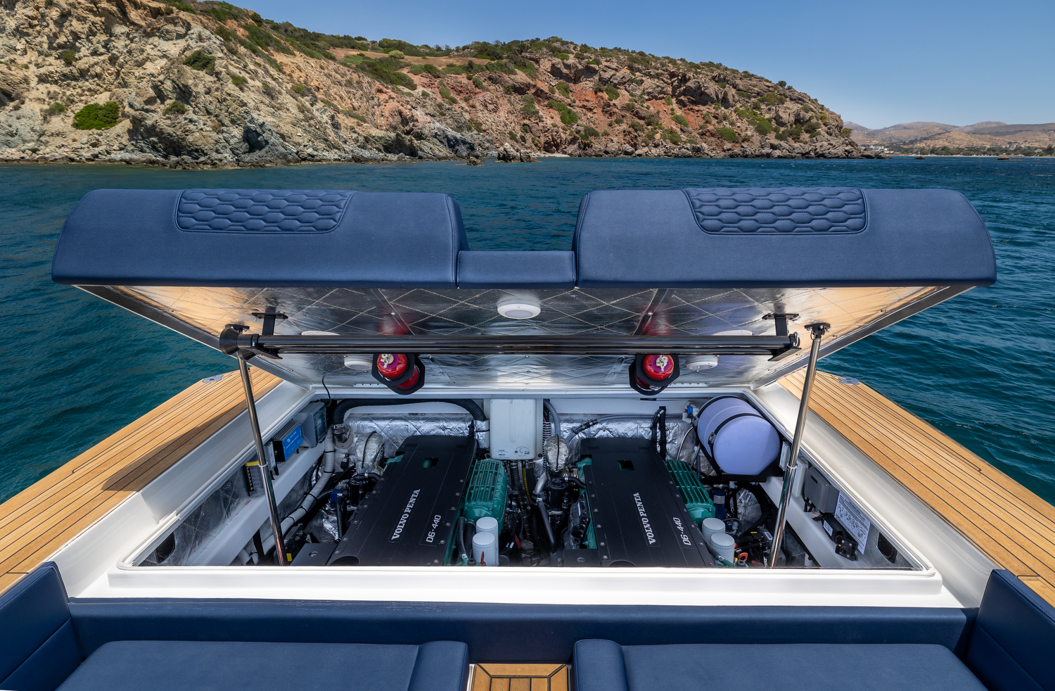 The engine bay open under the aft sunpad, showing two Volvo Penta D6-440 inboard diesels side by side beneath the lifted cushion, with a rocky headland behind