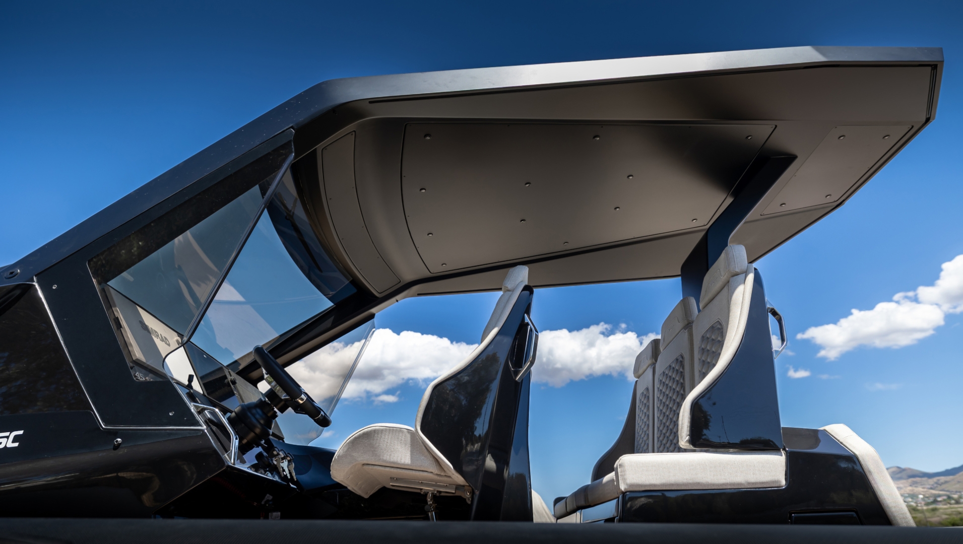The hardtop seen from below and behind, its dark underside spanning two white bolstered helm seats and the top of the windscreen, against a blue sky