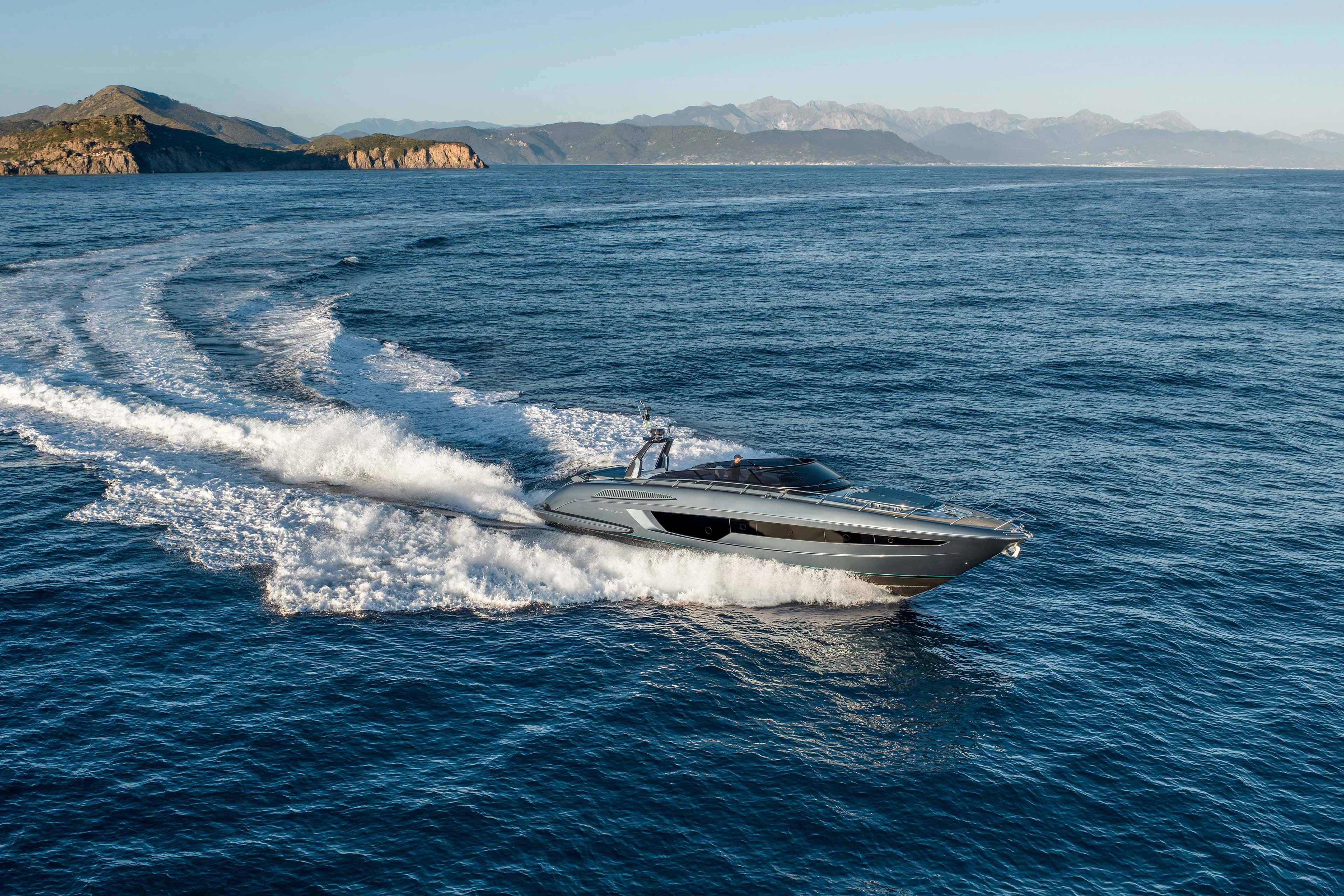 The yacht running away from the camera in a long curve, her wake sweeping across deep blue water towards a mountainous coastline