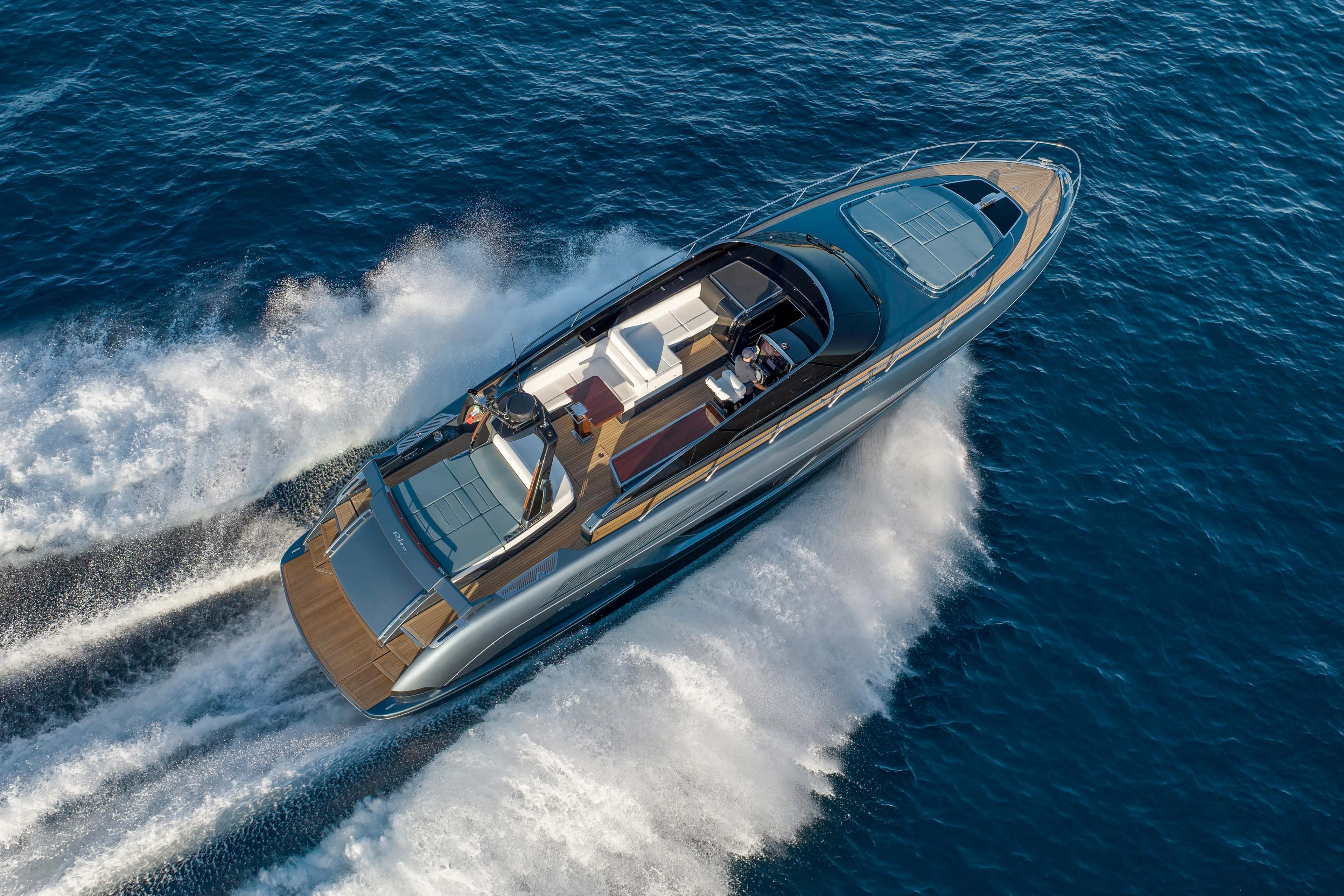 The yacht seen from above and astern at speed, the whole cockpit laid open with the helm, sunpads and a folded table on teak, and twin wakes spreading behind
