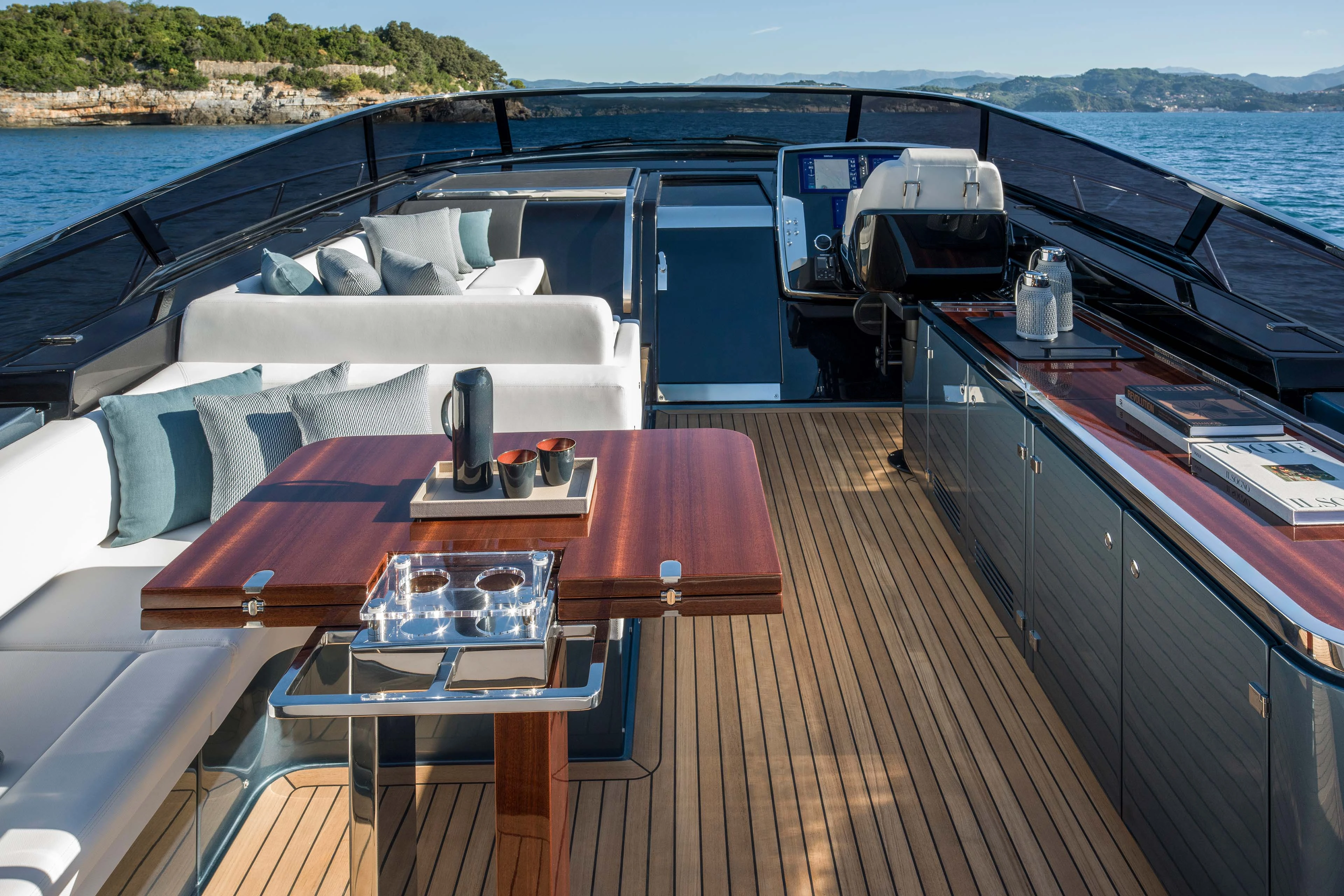 The cockpit looking forward, a polished wooden table with a drinks tray set between an L-shaped white sofa and a bar unit, with the helm and its white seats beyond