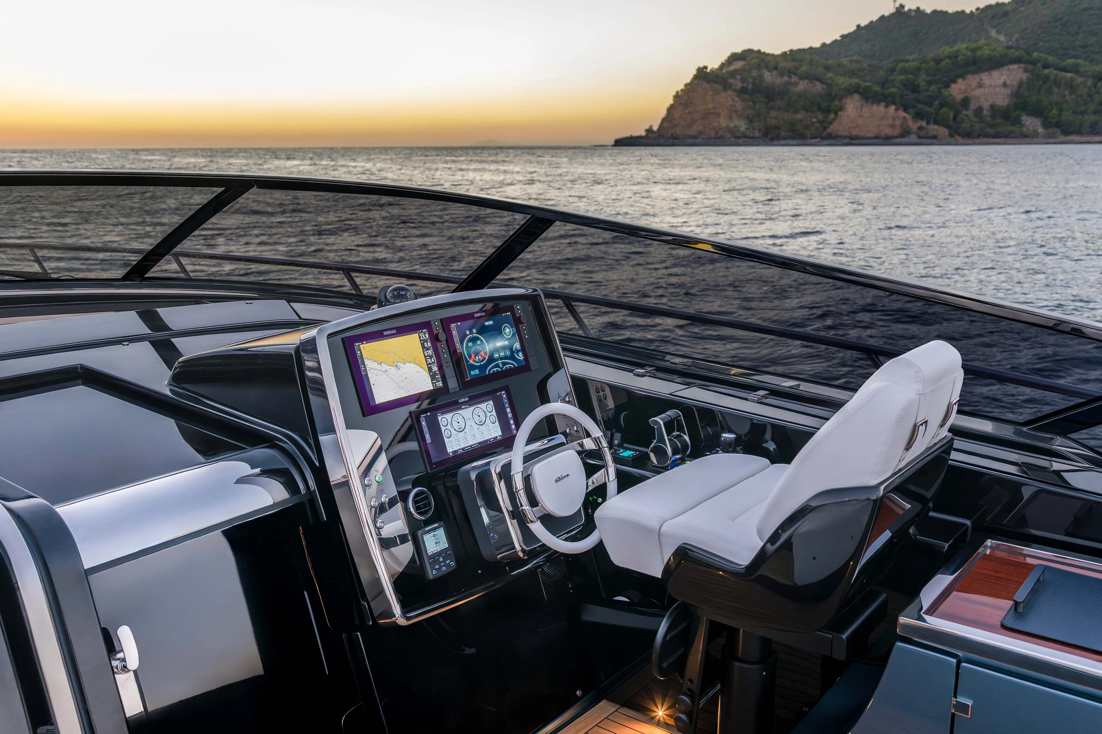 The helm at sunset, twin Simrad screens and a white-rimmed wheel set into a polished console with a single white helm seat beside it and a headland beyond