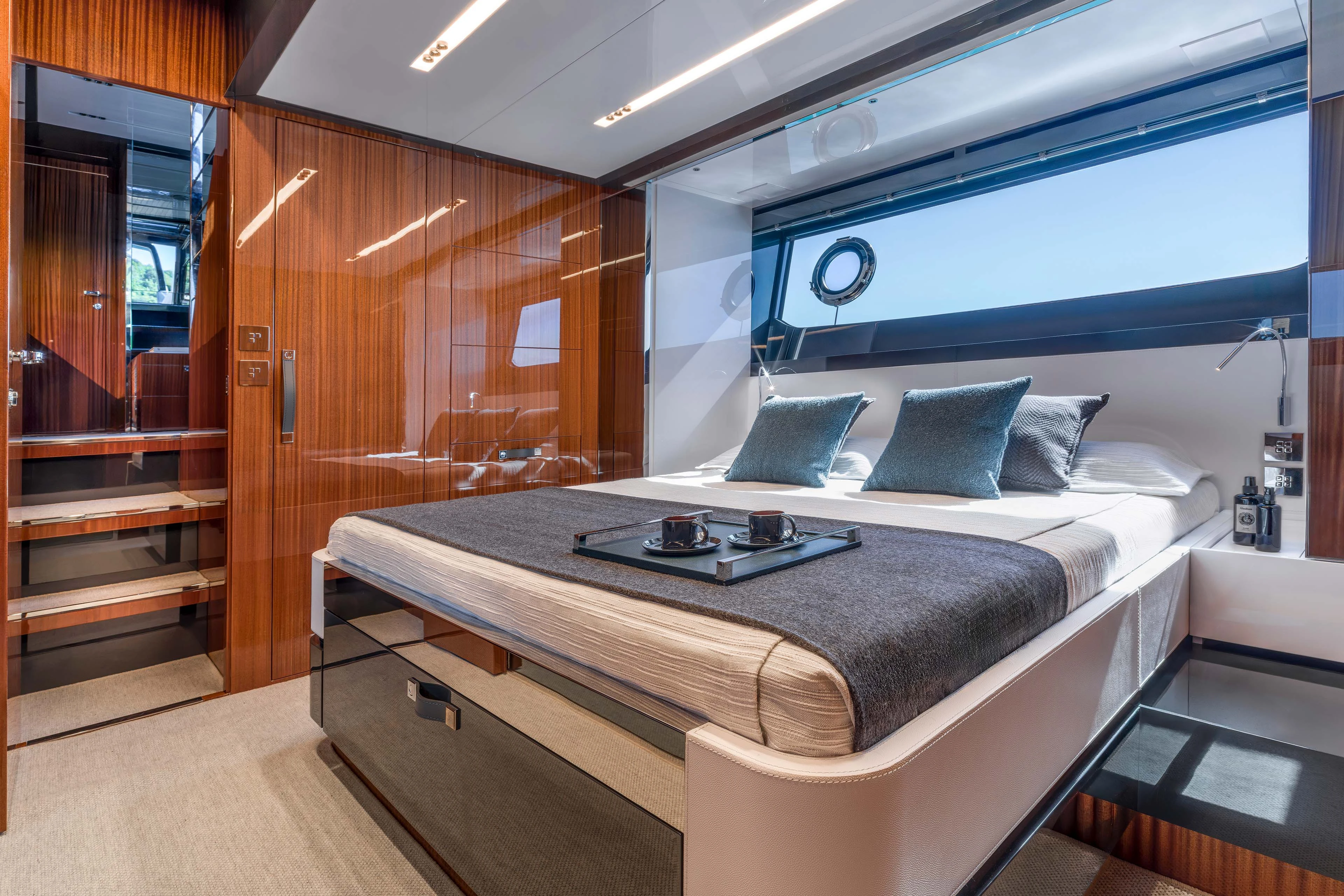 A double cabin with the bed against polished mahogany joinery, a long narrow hull window with a porthole above it and a breakfast tray set on the bedcover