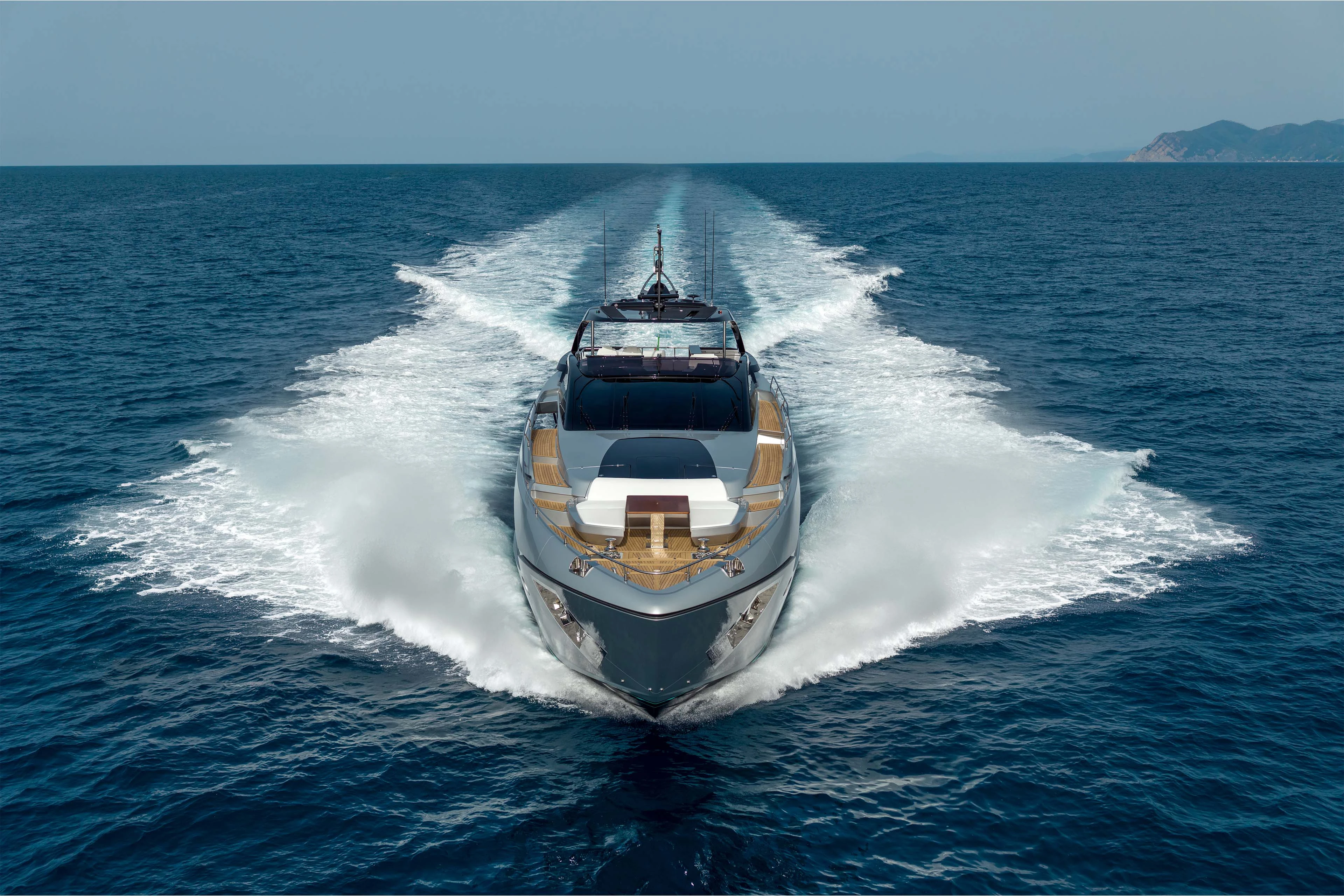 The Riva 96 Argo Super seen head on at speed, her grey hull parting the water and twin wakes fanning out behind her towards a distant headland