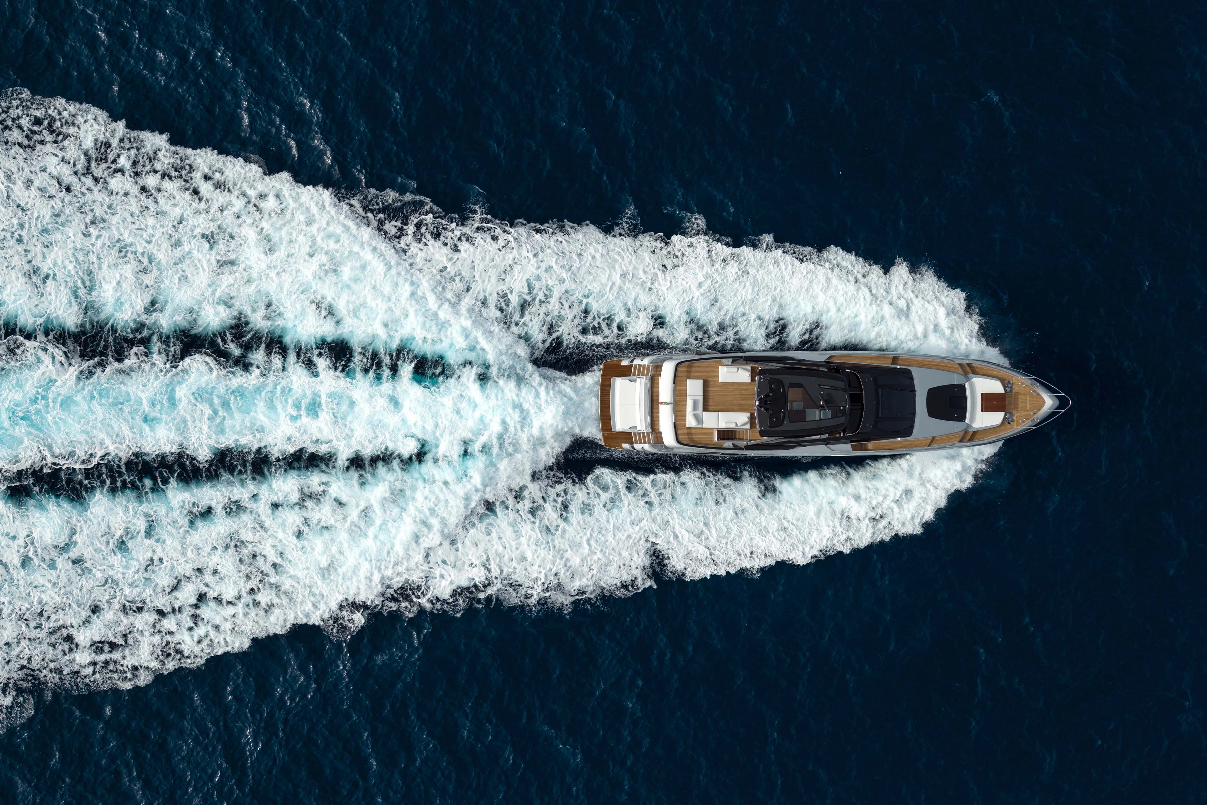 The yacht photographed from directly overhead at speed, teak decks and white sunpads laid out along her length between long white wakes