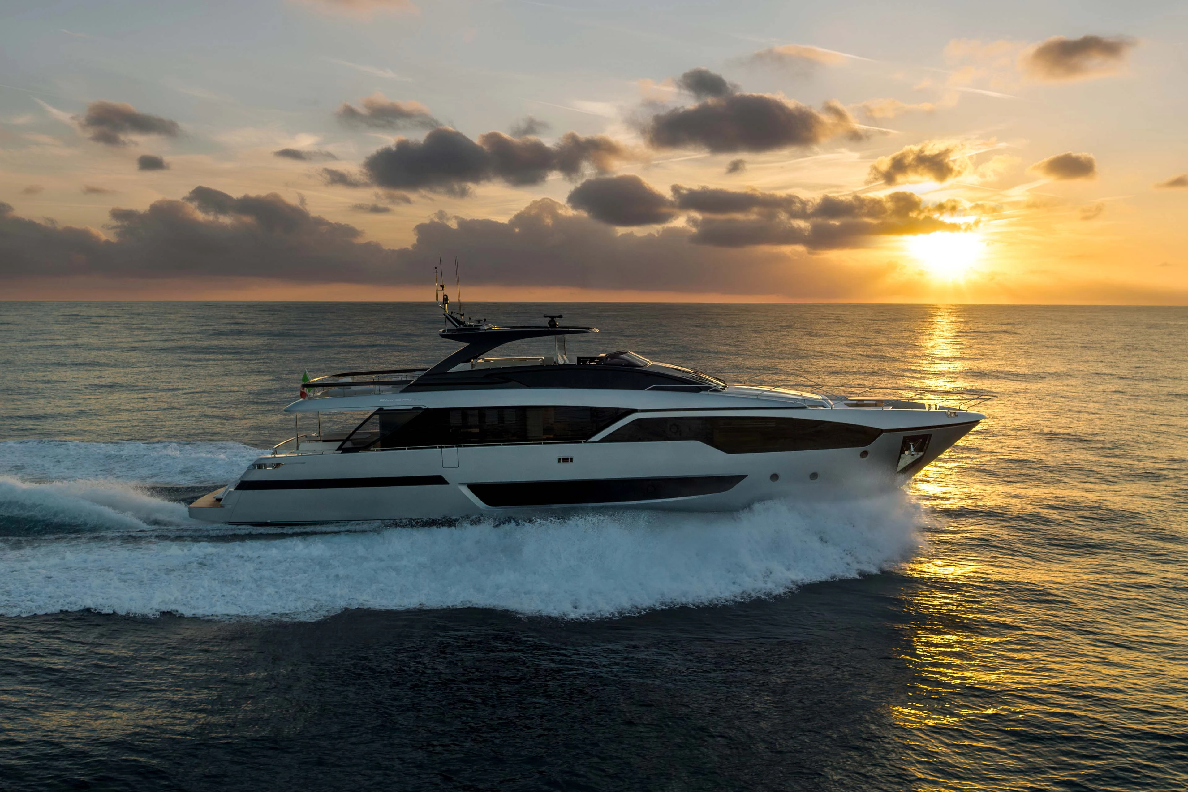 The yacht running left to right into a low sun, her profile dark against the water with the flybridge arch silhouetted