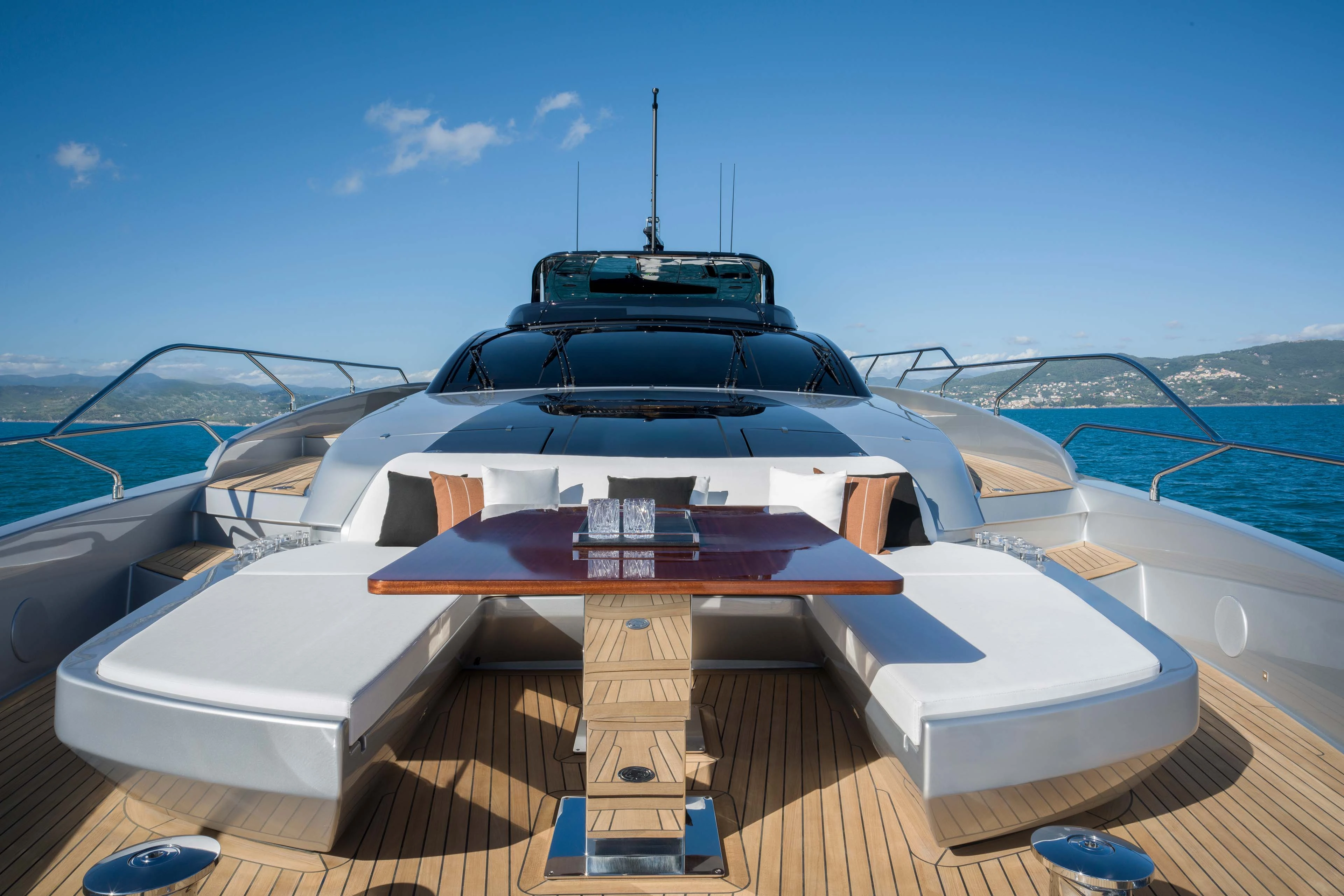 The foredeck seating seen from the bow: a large sunpad either side of a polished wooden table set on teak, with the windscreen and dark hardtop rising behind and a green coastline astern