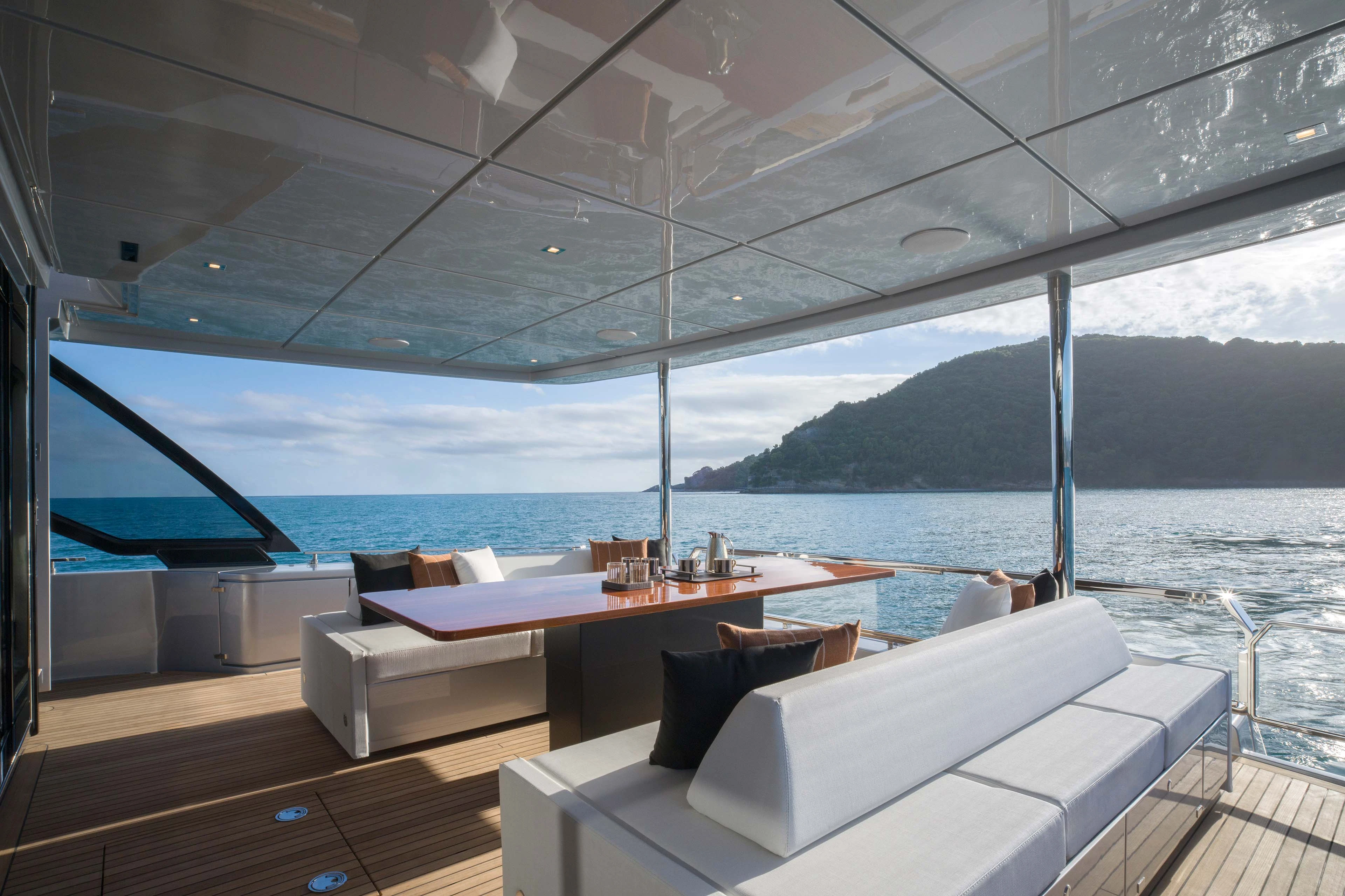 The aft deck under its mirrored overhead, a long wooden dining table with bench seating and a white sofa facing out over the open stern to a wooded headland