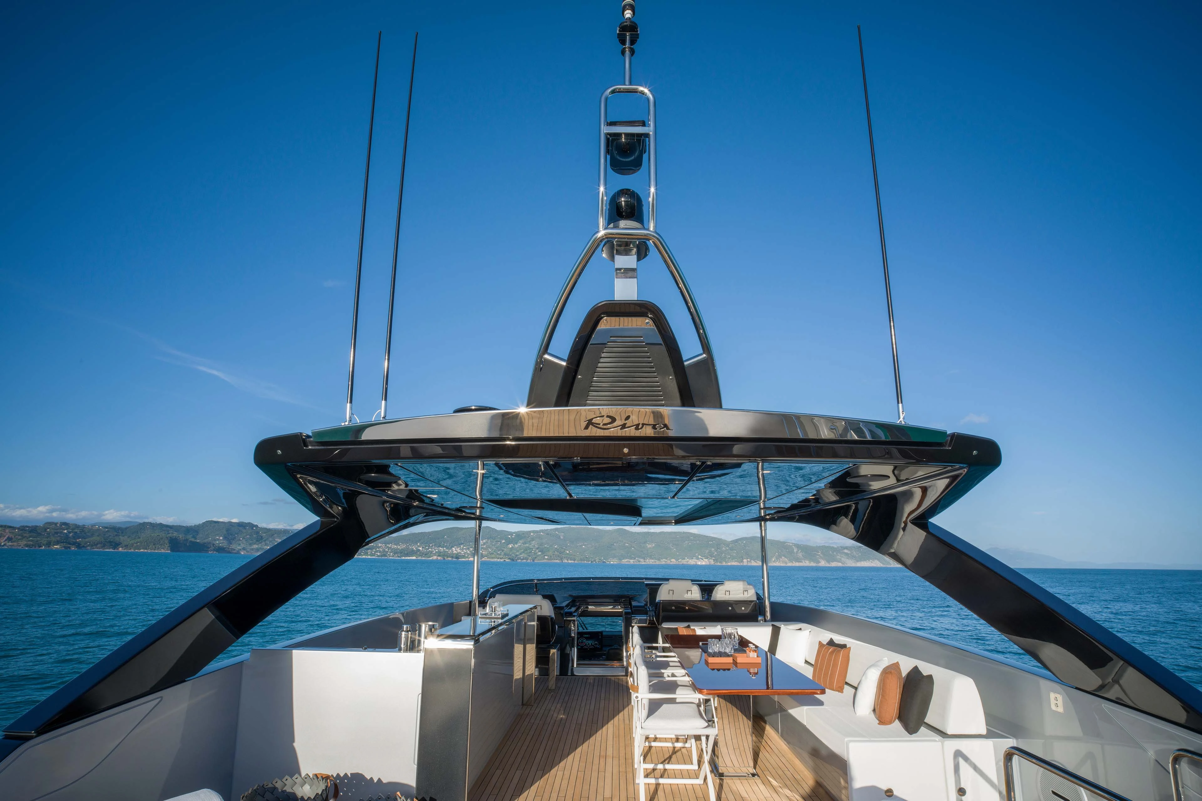 The sun deck seen from forward, the polished dark arch carrying the Riva name spanning the deck above the helm seats, bar and dining table, with the coast beyond