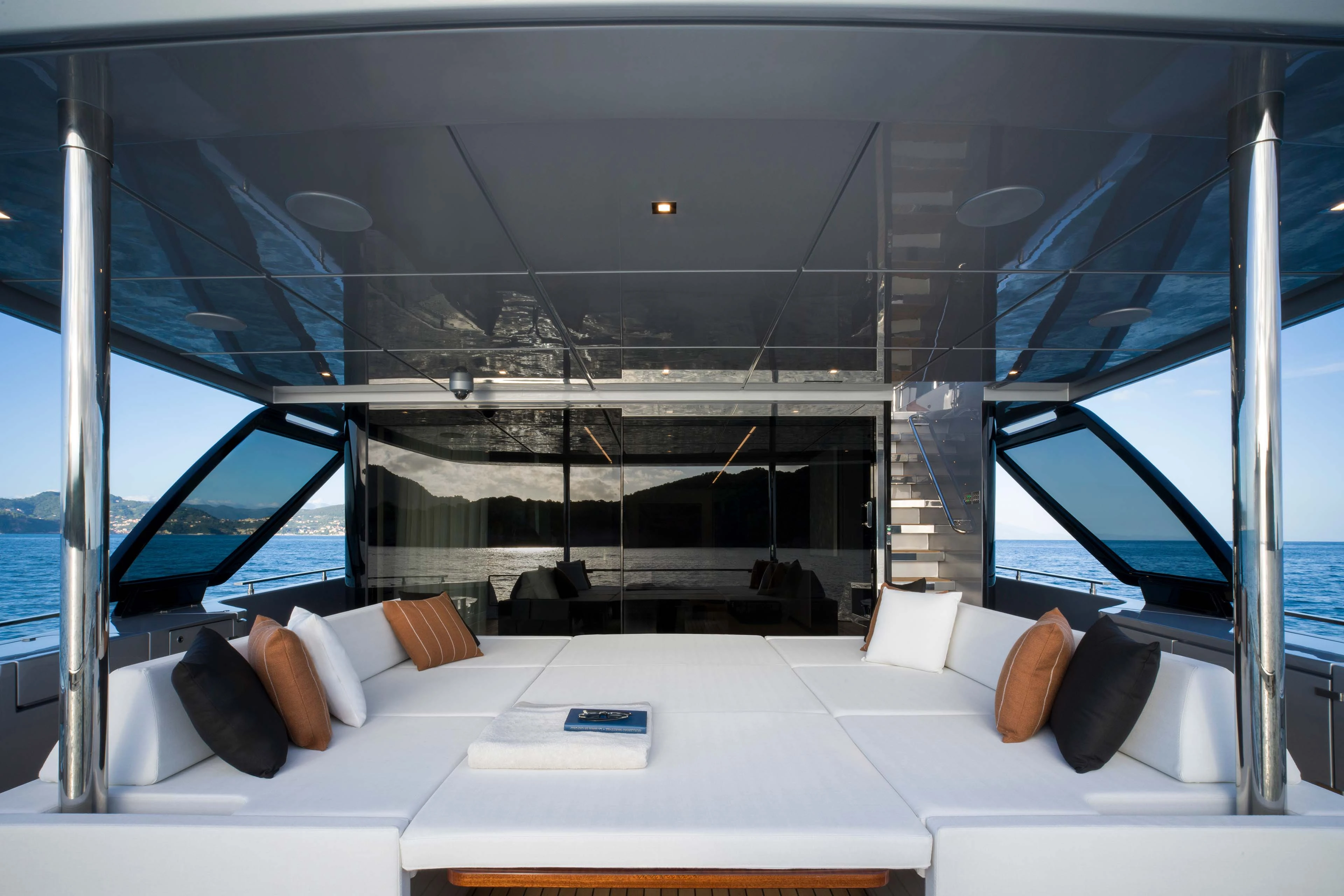 A wide sunbed filling the aft deck under a dark tinted overhead, with bolster cushions along both sides and the glazed saloon doors and a staircase beyond