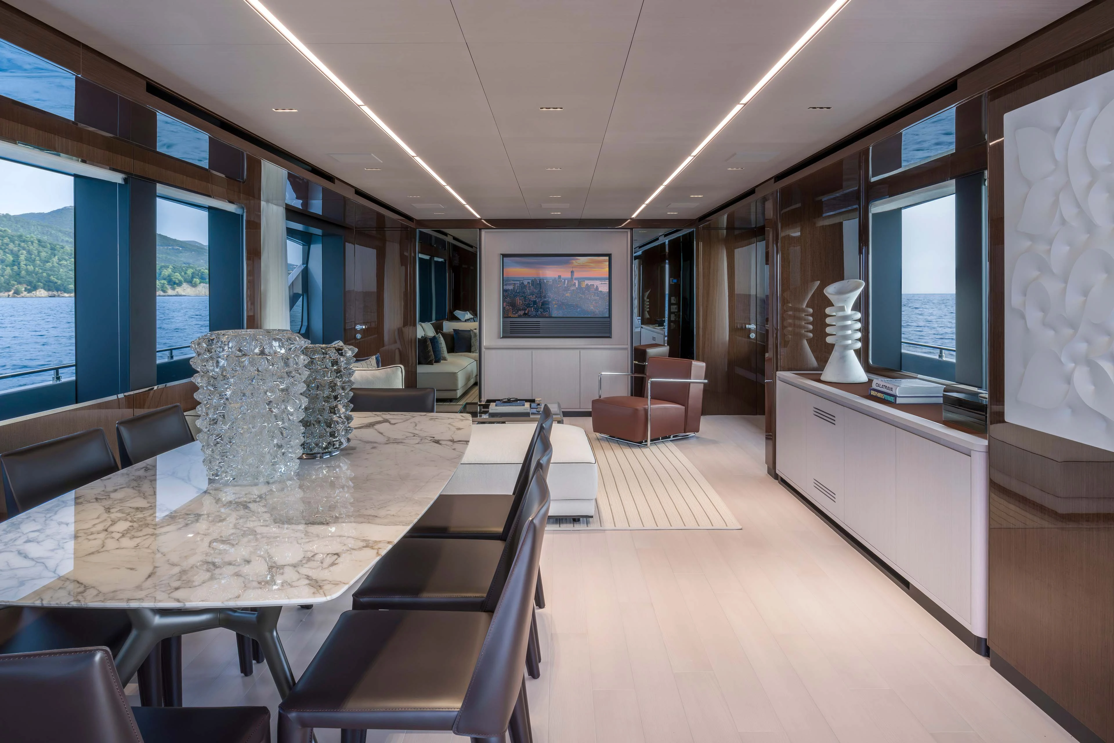 The saloon looking aft, a marble dining table with dark leather chairs in the foreground and a seating area with a leather armchair and television beyond, windows down both sides