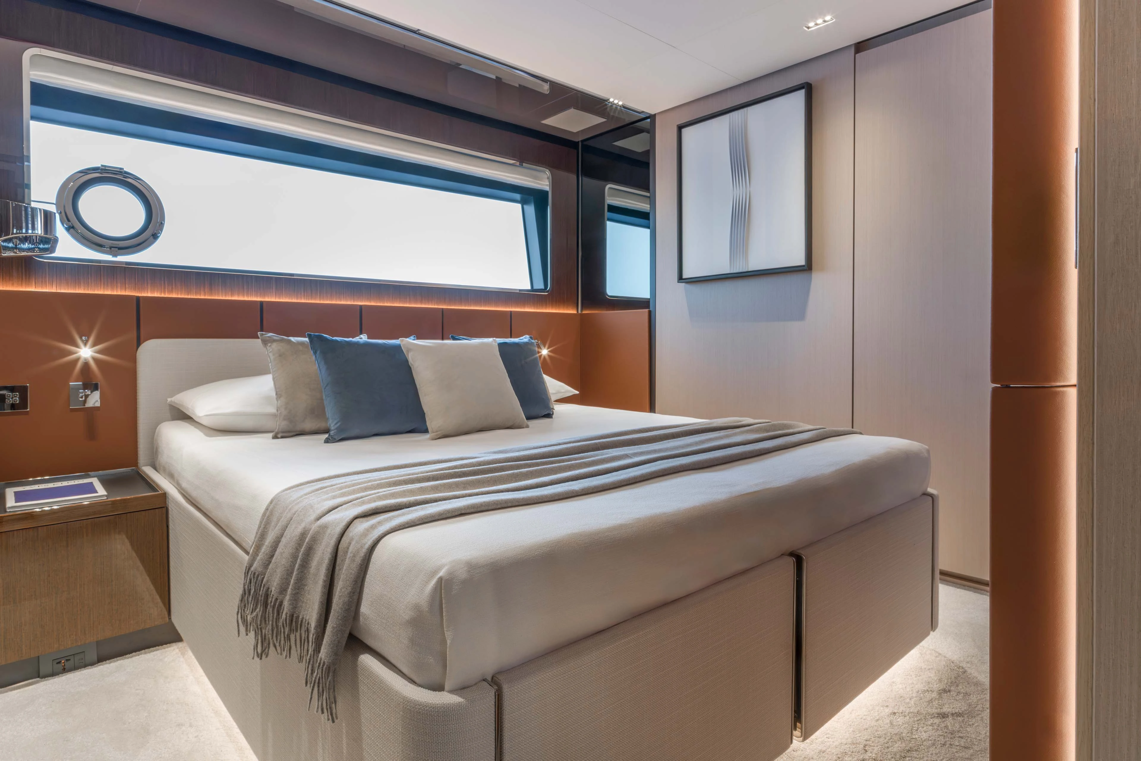 A guest cabin with a double bed against a tan headboard, a long narrow hull window with a porthole beside it and a framed print on the facing wall