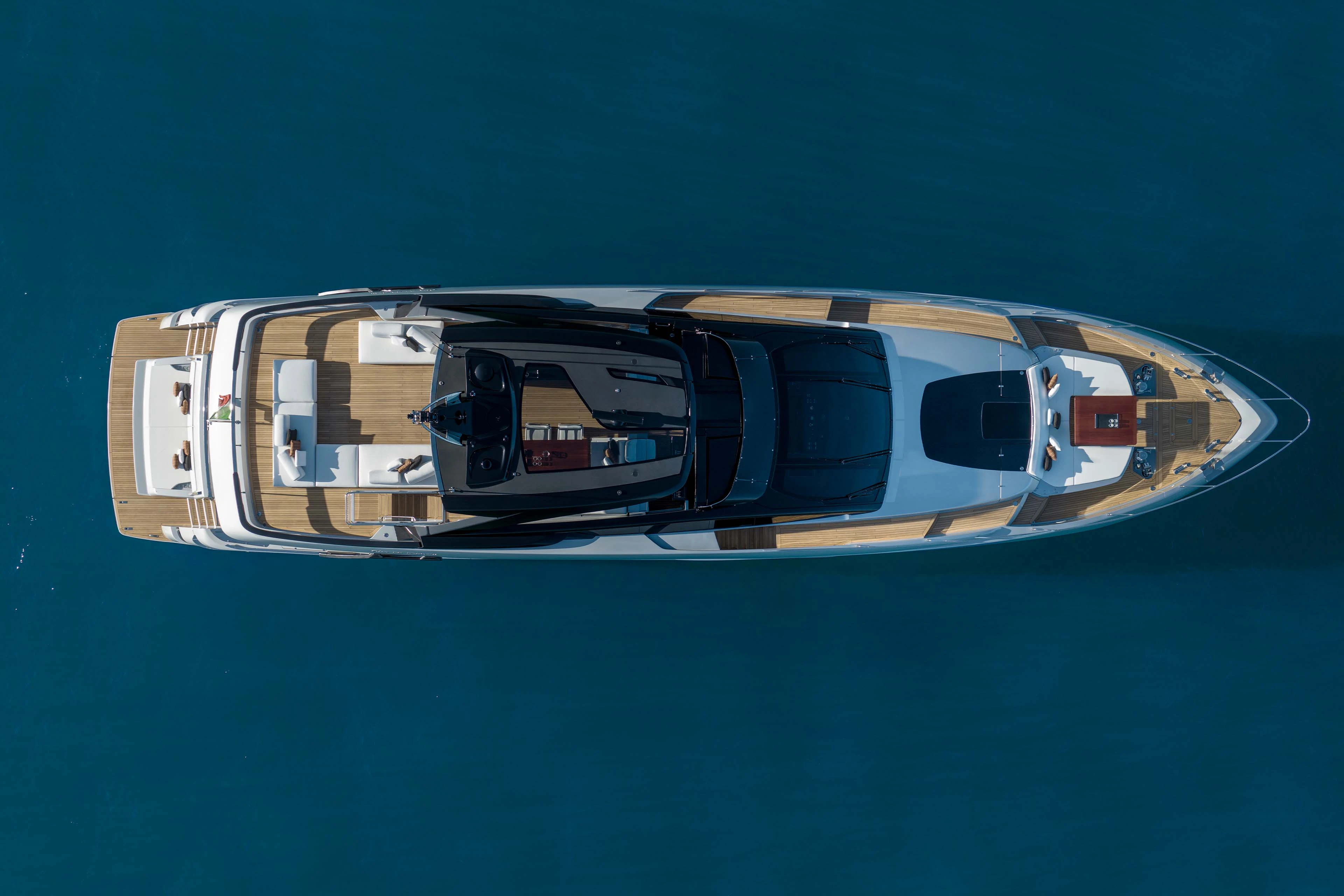 The yacht seen from directly overhead lying still on dark blue water, showing the teak side decks, the open sun deck and the tender stowed forward