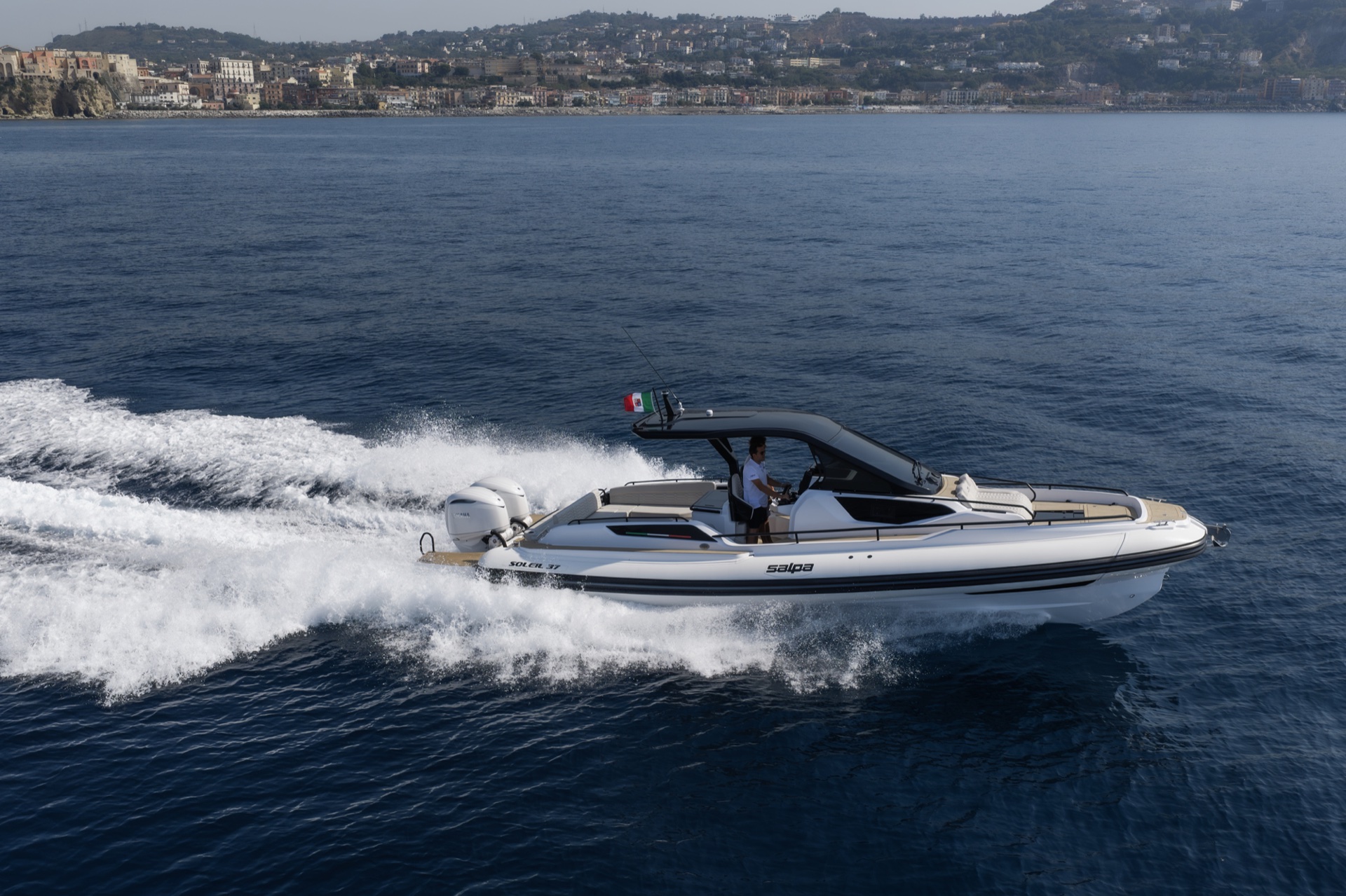 The Soleil 37 running across the frame with its wake behind, an Italian flag on the T-top and a hillside town beyond
