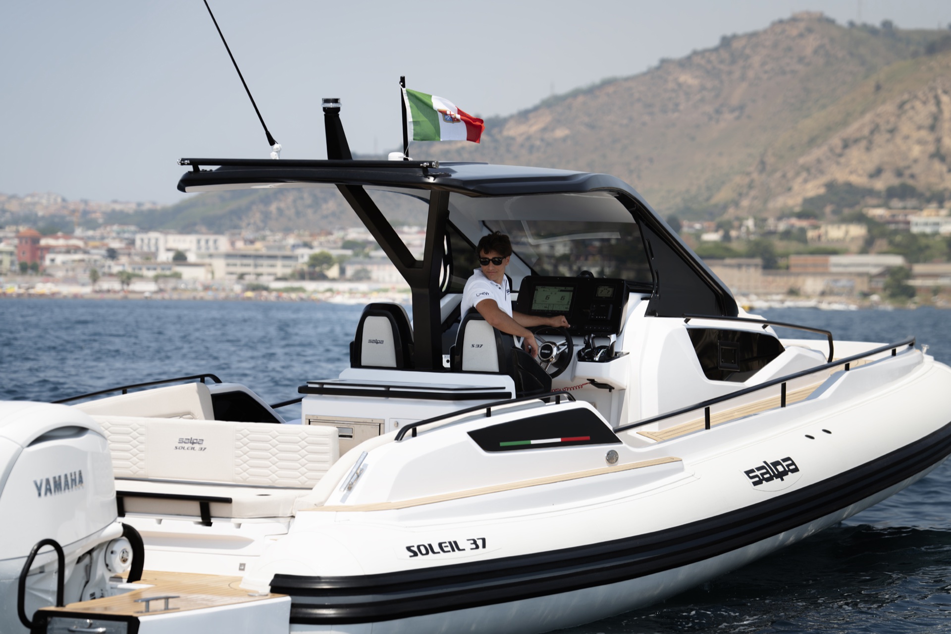 The helm of the Soleil 37 from the quarter, a driver at the wheel beneath the T-top, SOLEIL 37 lettered on the tube