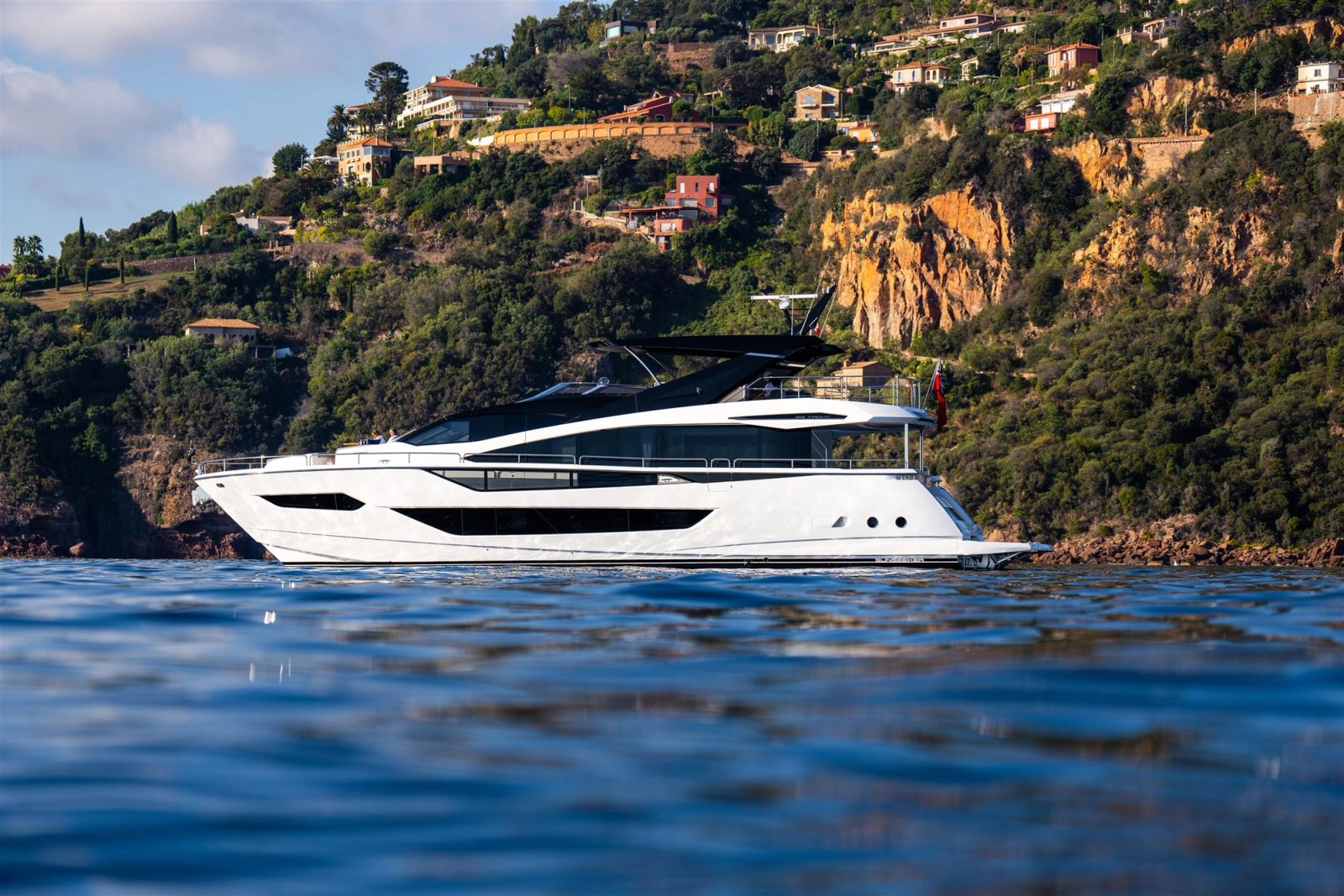 The Manhattan 94 lying still close inshore below a wooded hillside of villas on the French Riviera, seen exactly side on from water level, a red ensign at her stern