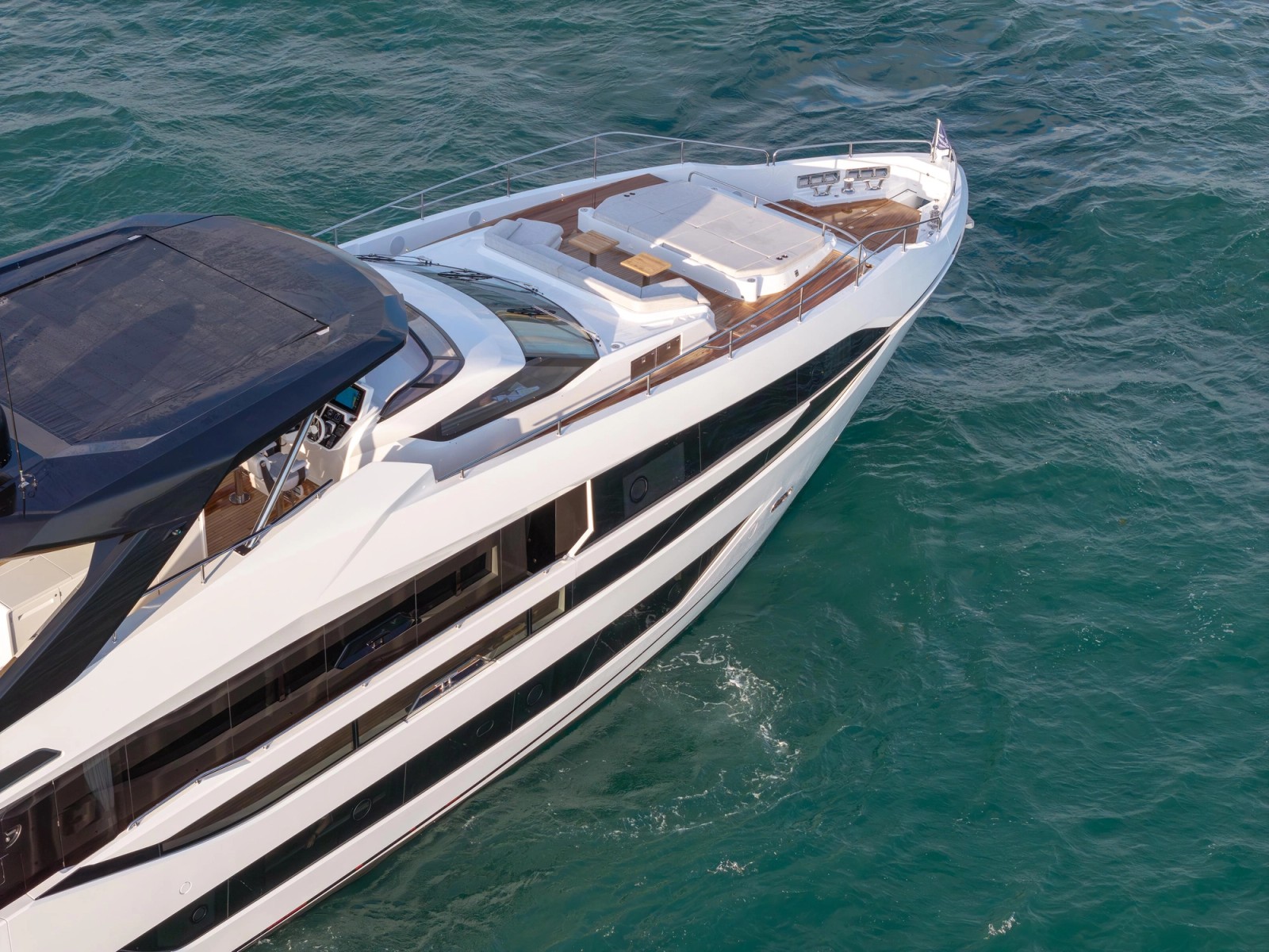 Looking down on the foredeck from above: a large sunpad and two separate seating areas in white upholstery set into teak, with the dark flybridge hardtop at the near edge of the frame