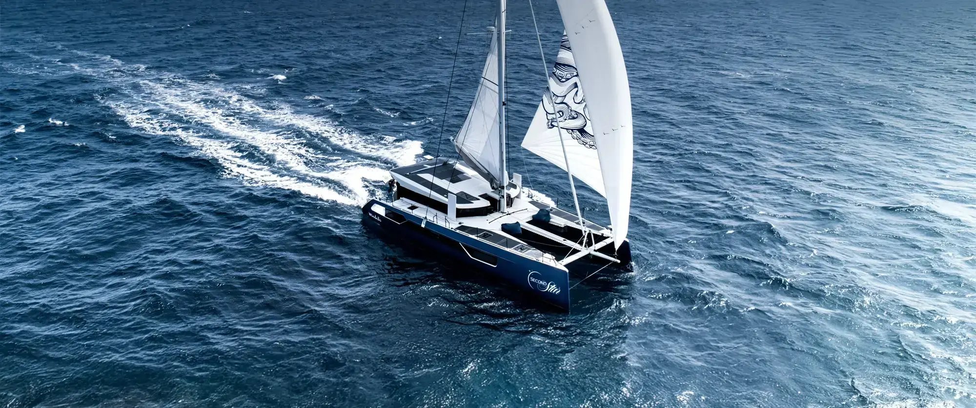 The catamaran seen from high above under sail, both hulls cutting a wake