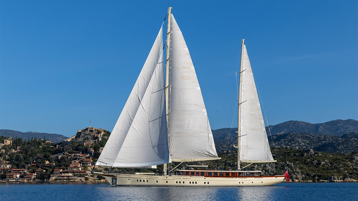 Simena broadside under full ketch rig, her name on the hull, with a hillside village and castle behind