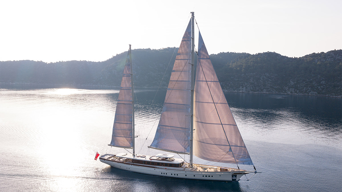 Simena photographed from above in backlight, both masts drawing, on flat water below a wooded shore