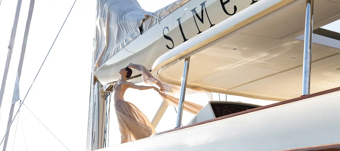 A photograph looking up at Simena's boom lettered SIMENA, a guest in a long dress on the side deck below the furled mainsail