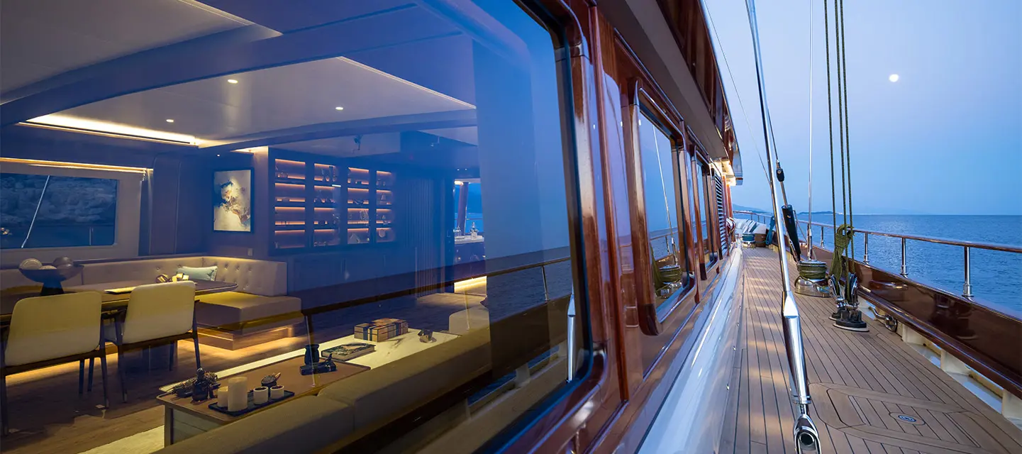 A photograph along the side deck at dusk, the lit saloon visible through long windows and mahogany-framed glazing, sea and moon beyond