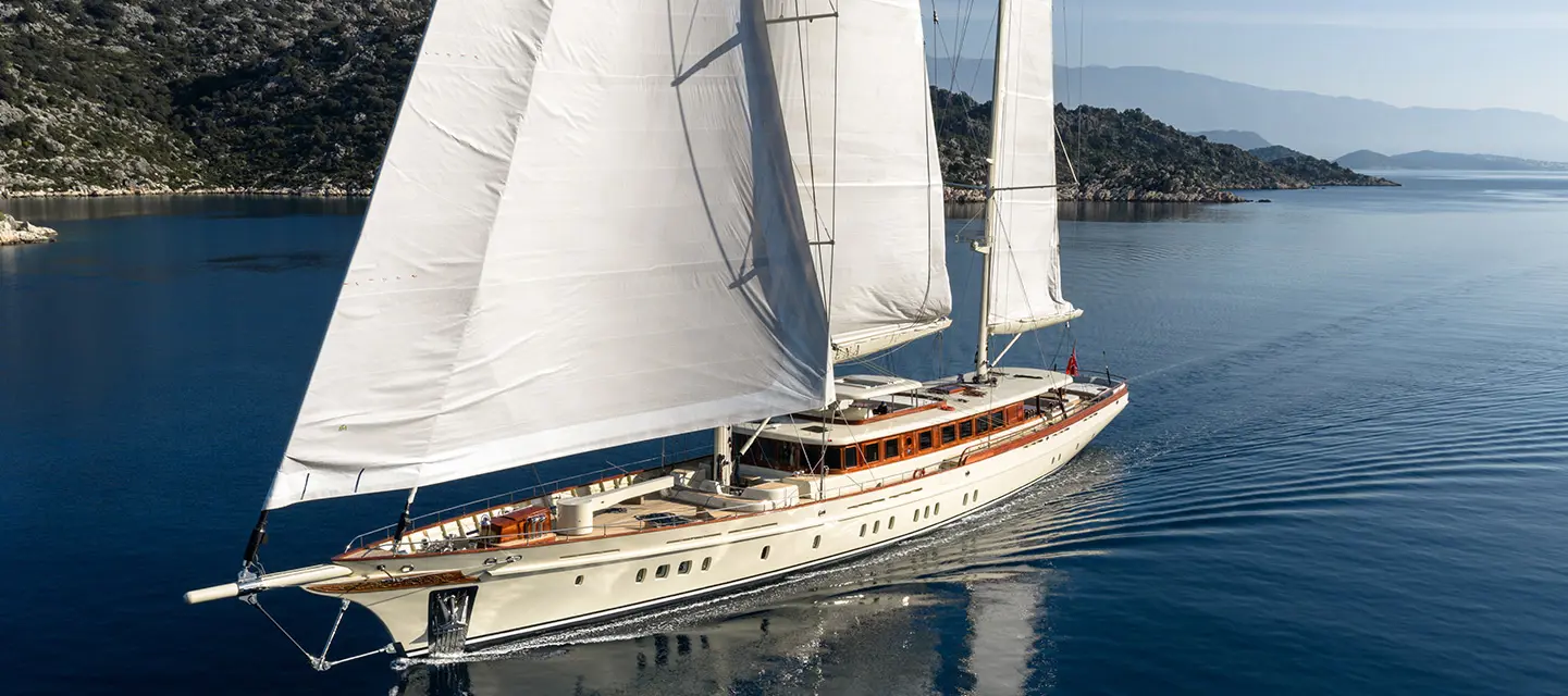 A photograph of the main saloon, a dining table to port and an L-shaped lounge to starboard under a walnut ceiling, doors open aft to the cockpit