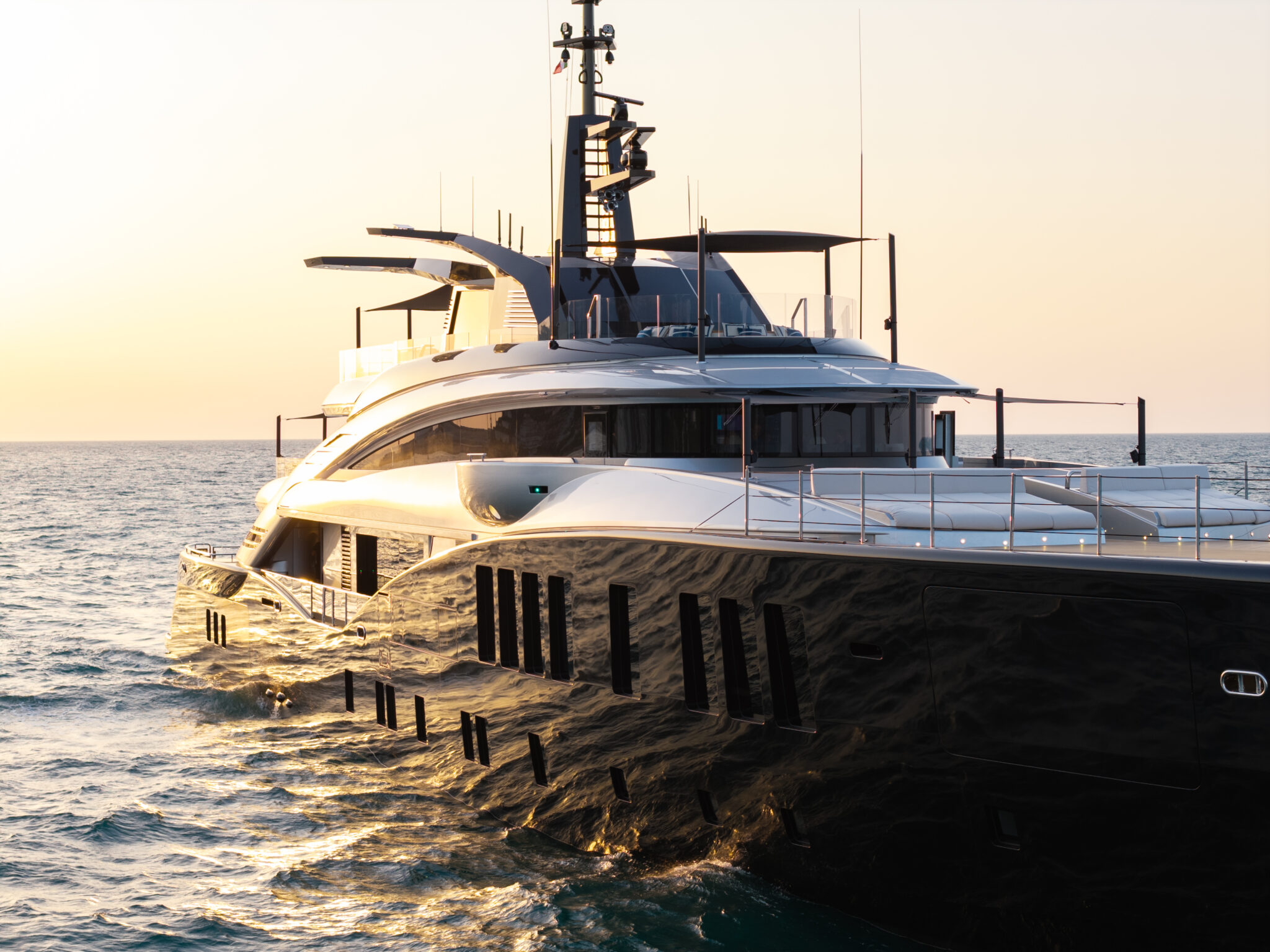 The yacht's aft quarter at sunset, showing the vertical window slots in the black hull and the sunpads on the deck above