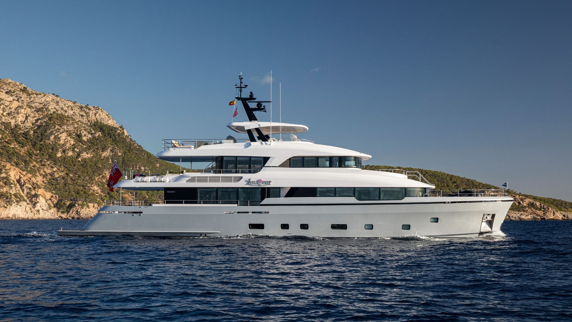 LOLLIPOP in full profile from her port side, three decks over a long silver hull, a sundeck hardtop and mast above, a rocky headland behind her
