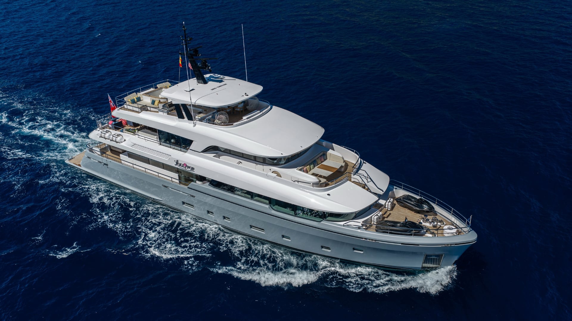 LOLLIPOP from above and astern on her port quarter, cutting a white wake across deep blue water, with sun loungers on the sundeck and tenders stowed on the foredeck