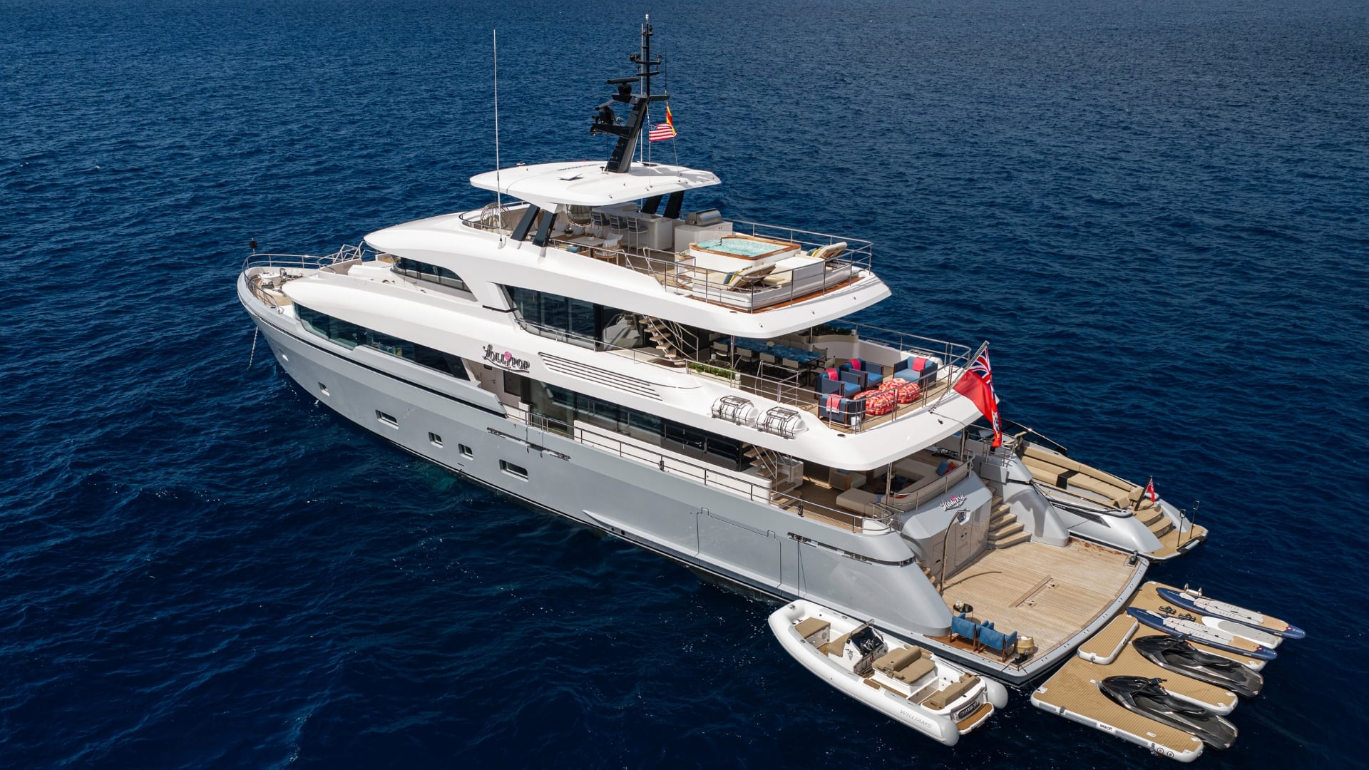 LOLLIPOP at anchor seen from above her starboard bow, the fold-out teak swim platform aft with a tender alongside and four jet skis on a floating dock, and a spa pool with sun loungers on the sundeck