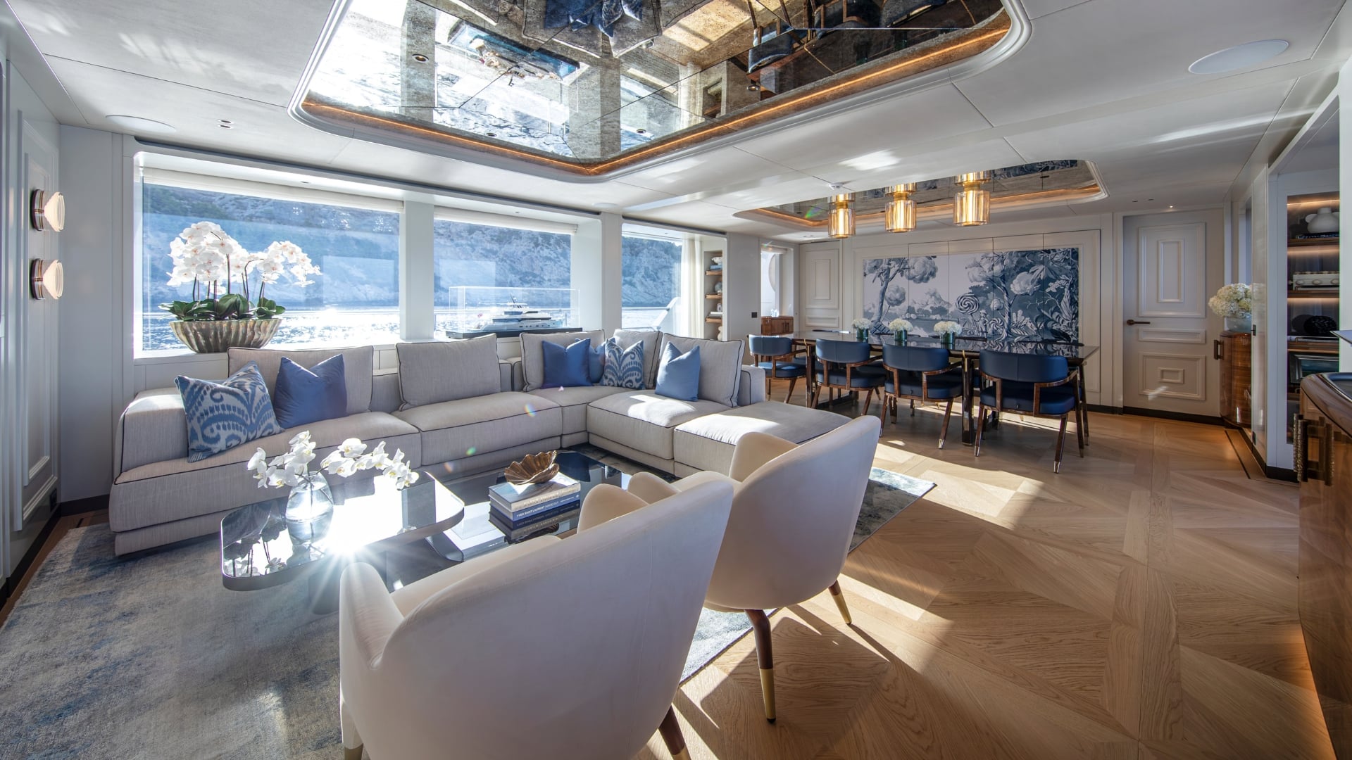 The main salon looking aft to the dining table, a pale corner sofa with blue cushions under full-height windows, a chevron oak floor and a mirrored ceiling panel above