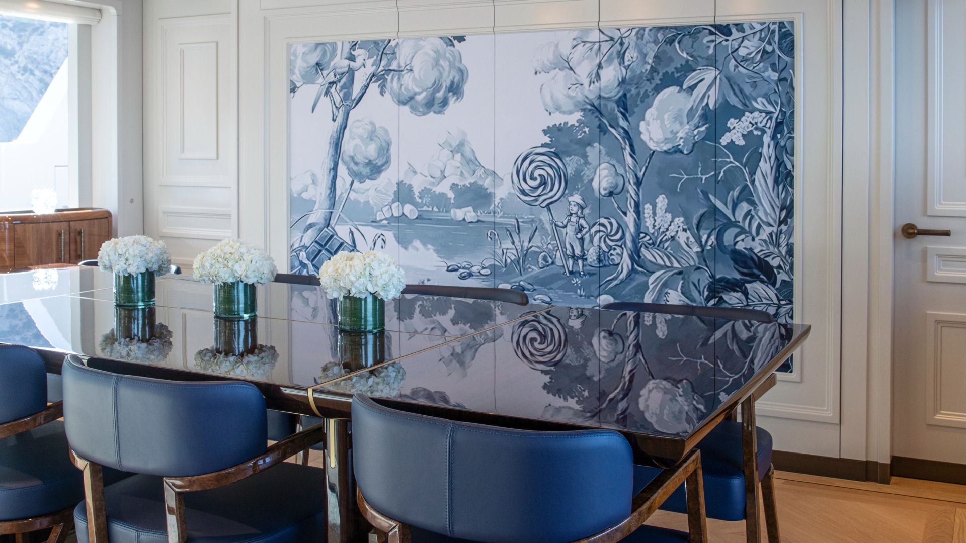 The dining table in dark polished wood with blue leather armchairs, set with white hydrangeas, in front of a wall-wide blue and white mural of a garden in which a child holds a giant swirled lollipop